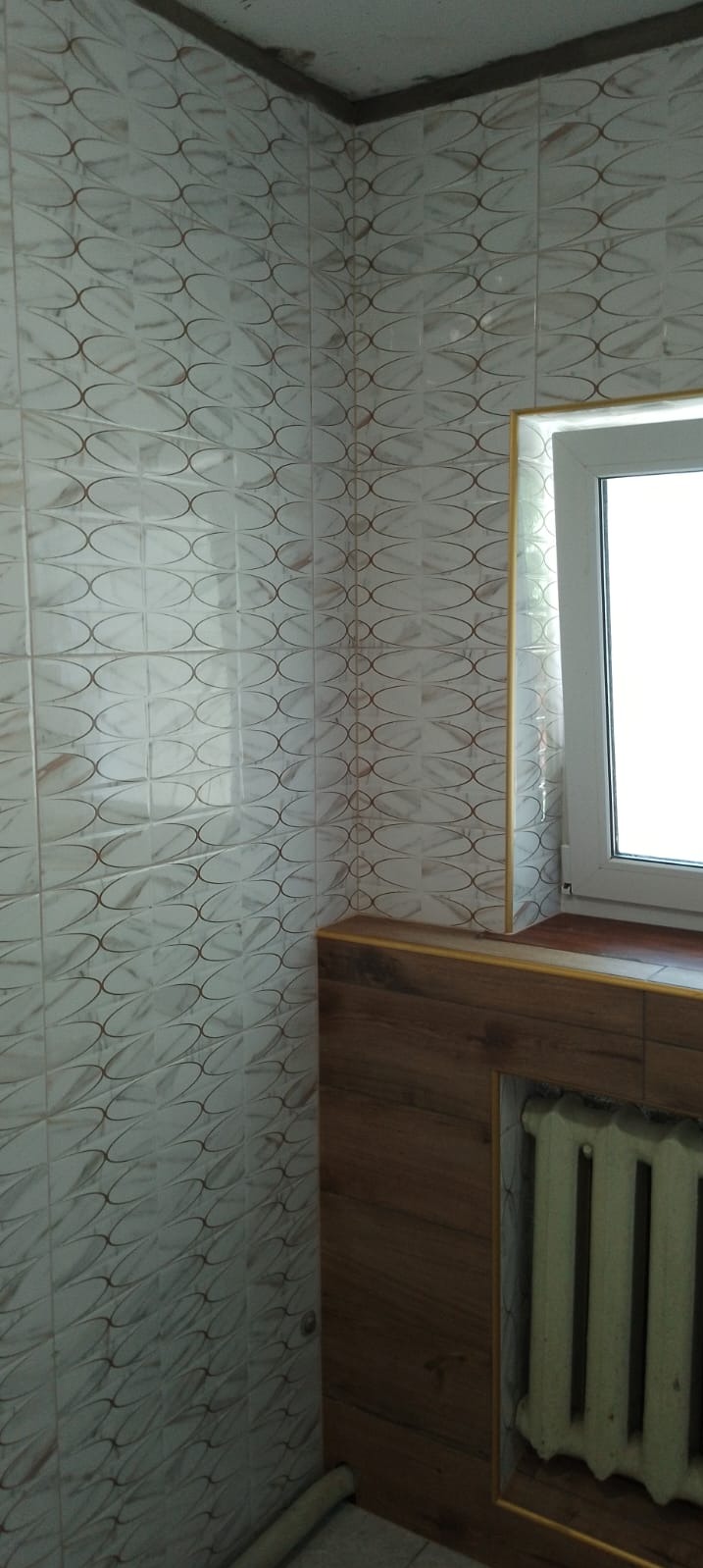 Wykończony narożnik łazienki z białymi płytkami w owalny wzór, drewnianą zabudową okna i grzejnikiem, widoczne niedociągnięcia w fugowaniu.
