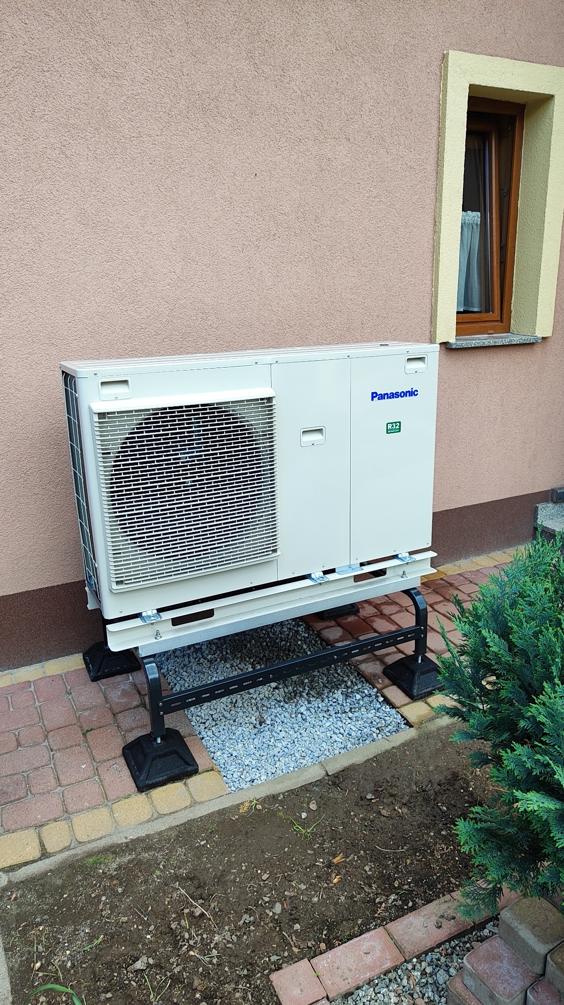 Zewnętrzna jednostka klimatyzacji Panasonic, zamontowana na metalowym stelażu z podkładem z drobnych kamieni, na tle elewacji budynku z oknem.