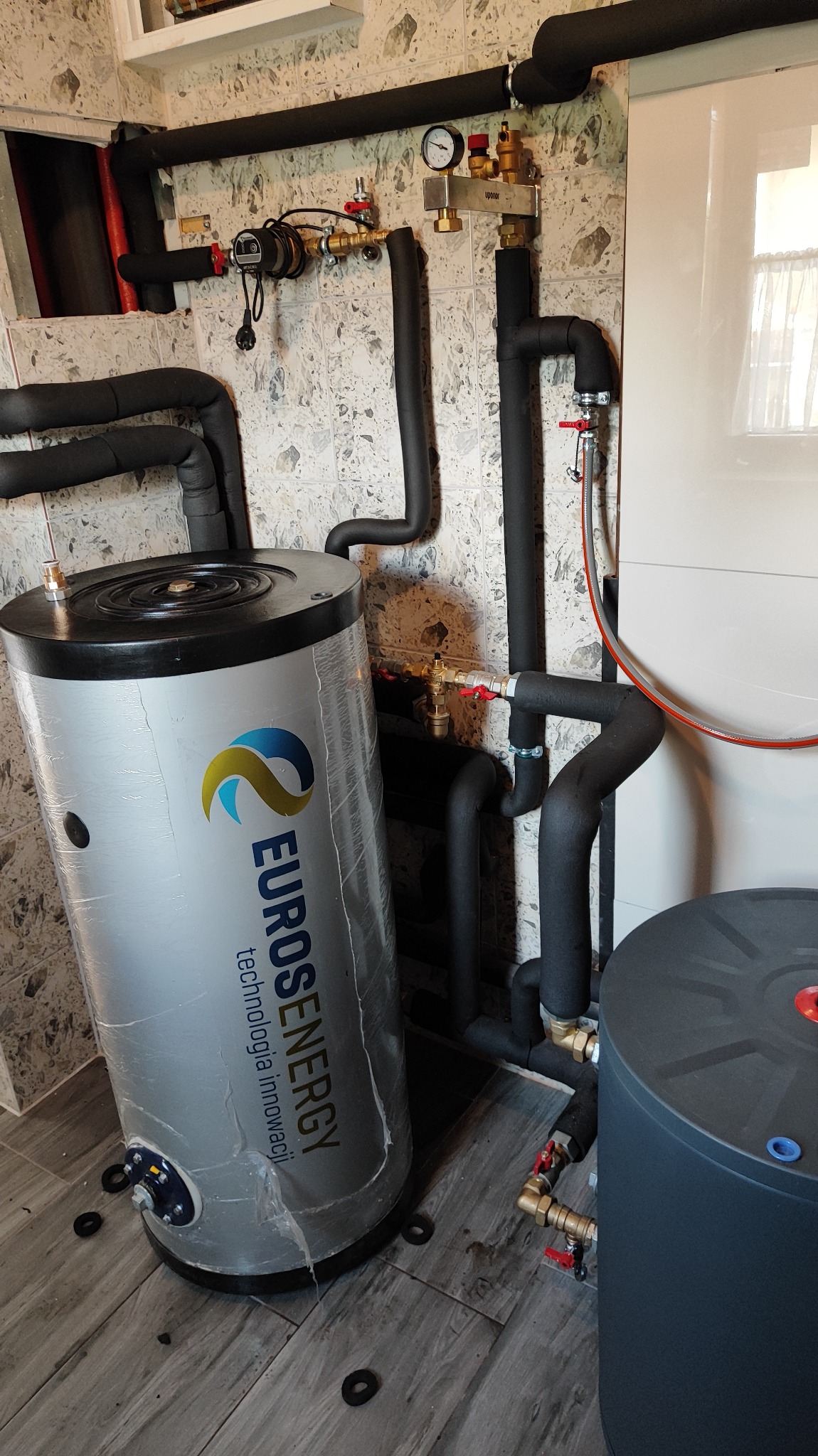 Szary, cylindryczny zbiornik z logo Euroenergy, obok ciemnoszarego zbiornika, z widocznymi rurami w izolacji, zaworami i manometrem na tle kafelkowanej ściany.