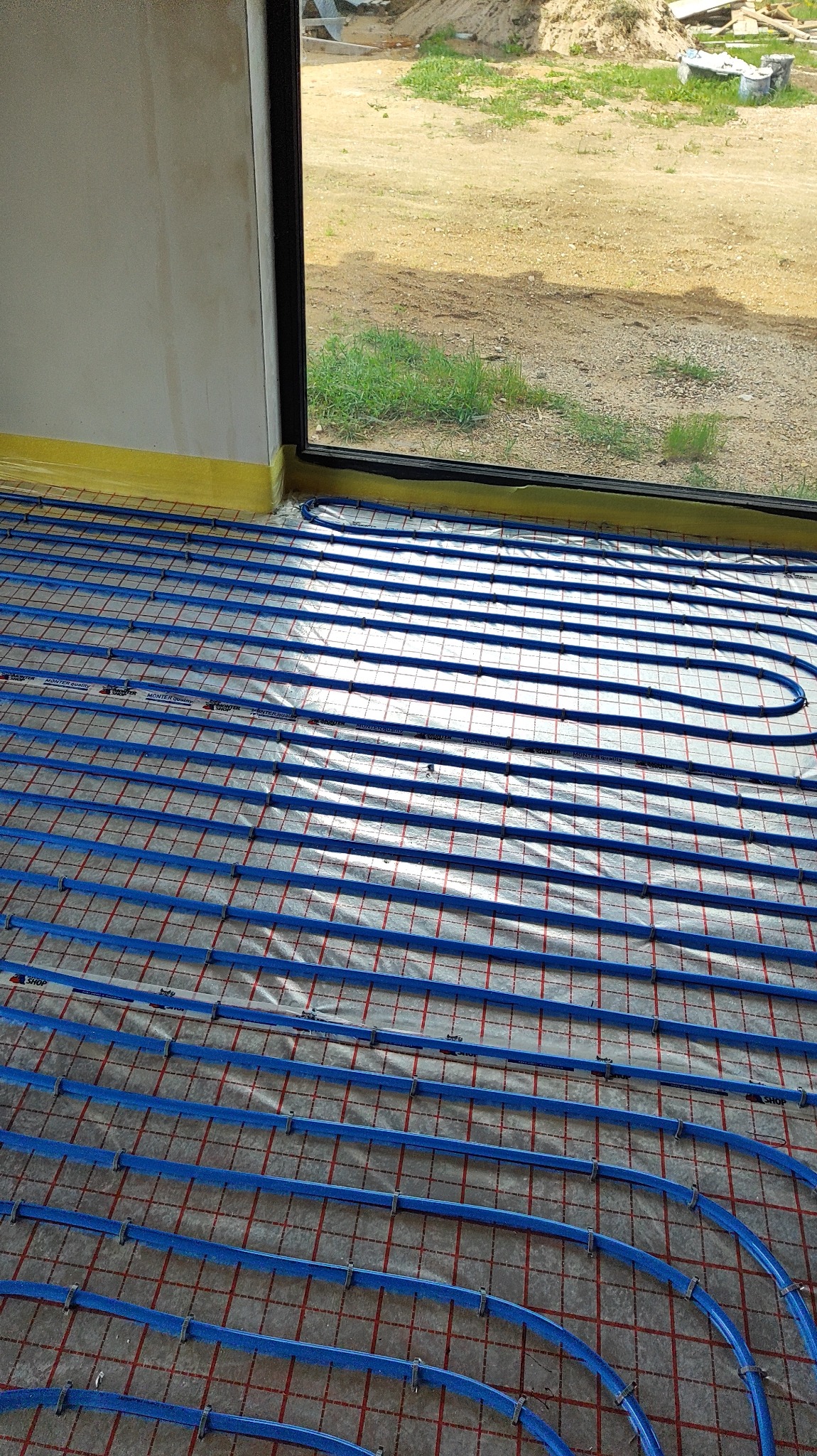 Niebieskie rury ogrzewania podłogowego, ułożone na folii z czerwoną siatką, widoczne przy ścianie z oknem na zewnątrz, zabezpieczone żółtą taśmą.