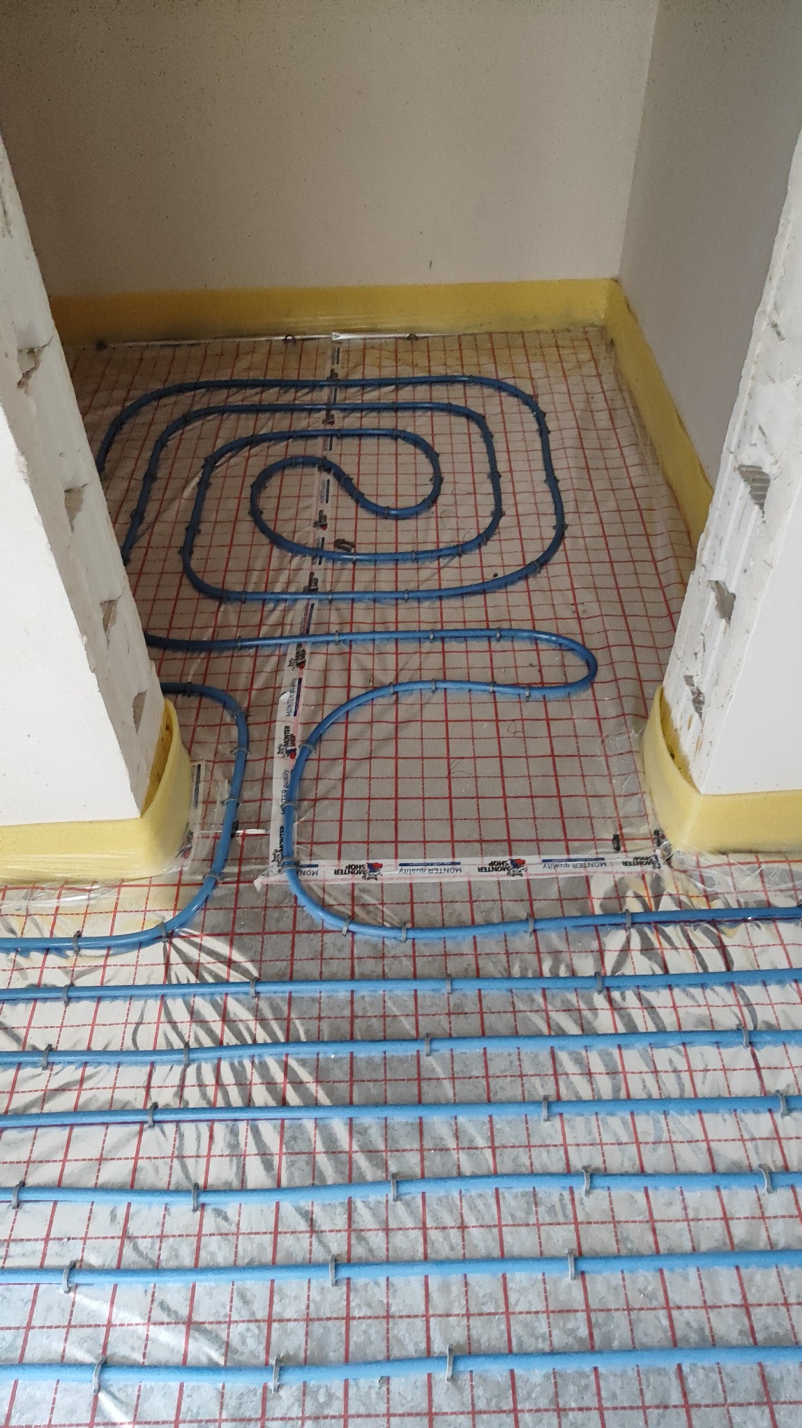 Niskie ujęcie instalacji ogrzewania podłogowego z niebieskich rurek ułożonych na folii z czerwoną siatką, widoczne fragmenty ścian z żółtą pianką dylatacyjną.