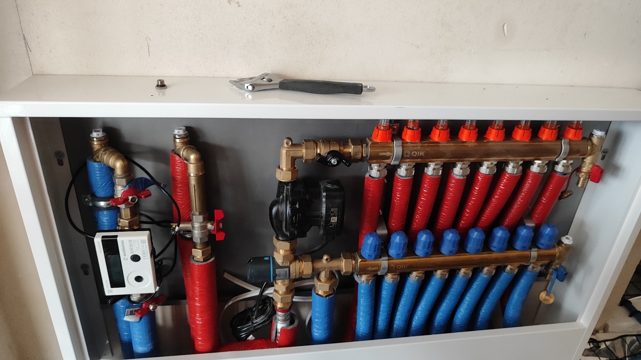 Rozdzielacz ogrzewania podłogowego z czerwonymi i niebieskimi rurami, umieszczony w białej szafce, z kluczem nastawnym leżącym na górze.