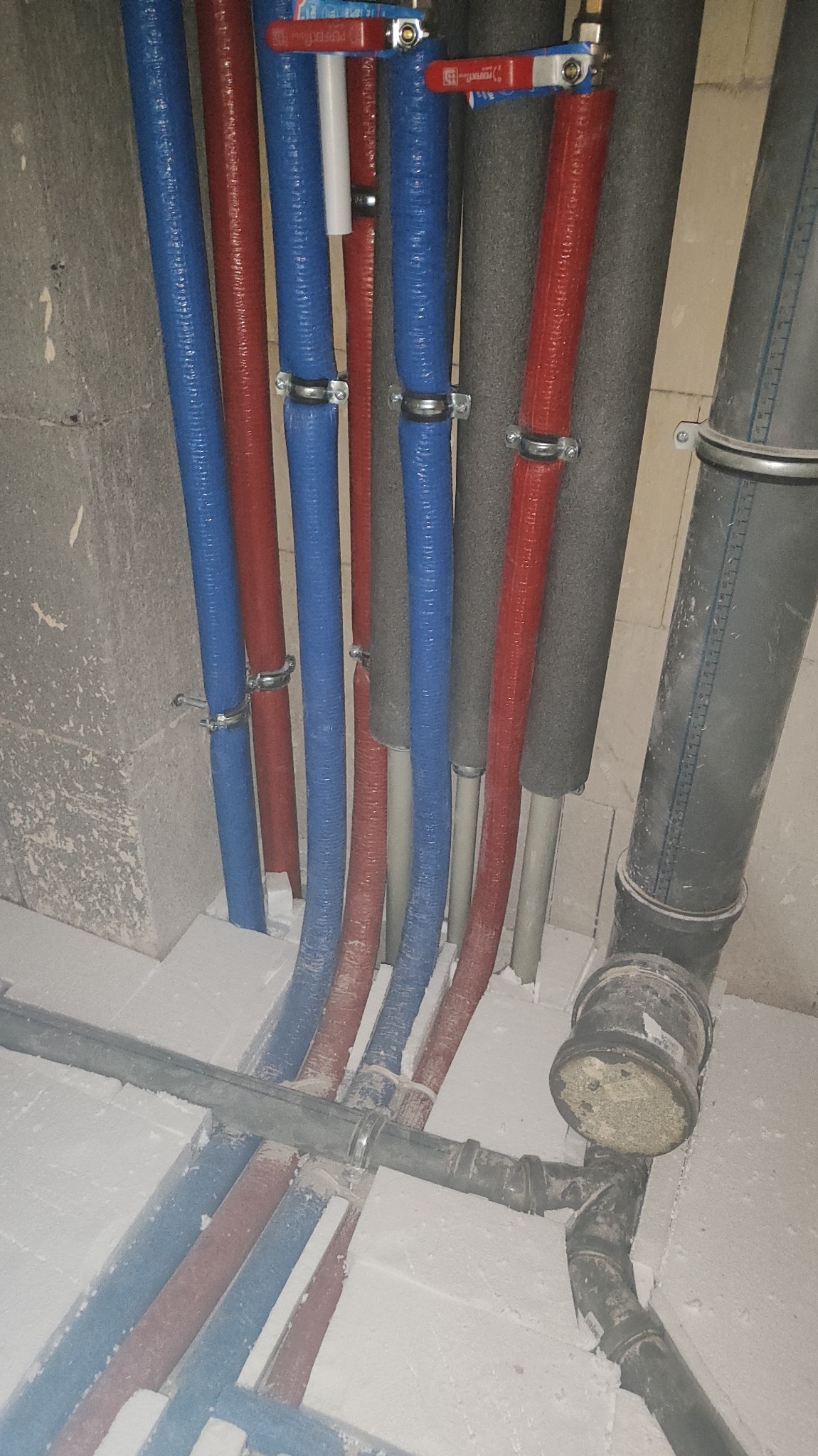 Rzędy niebieskich i czerwonych rur z zaworami, częściowo zaizolowanych, zamocowanych do ściany, wraz z fragmentem instalacji kanalizacyjnej w surowym stanie wykończenia.