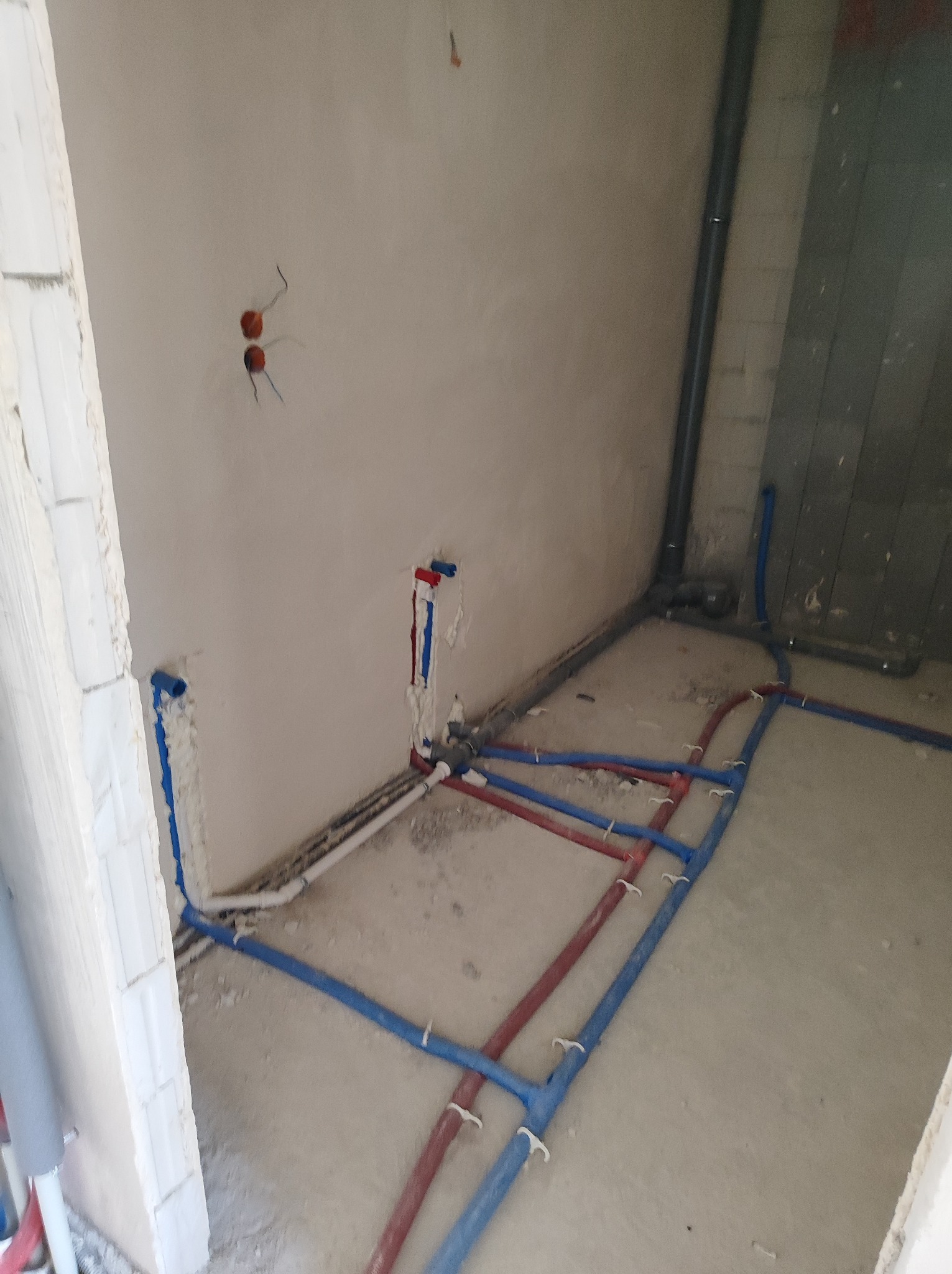 Surowe wnętrze w trakcie prac hydraulicznych: widoczne rury wodne w kolorach czerwonym i niebieskim, prowadzone po podłodze i ścianach, zabezpieczone pianką montażową w miejscach łączeń,...