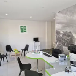 Jasne wnętrze placówki bankowej VeloBank z białymi ścianami, czarnymi krzesłami, biurkiem z zielonym akcentem i sprzętem biurowym, w tle fototapeta z czarno-białym motywem miejskim.