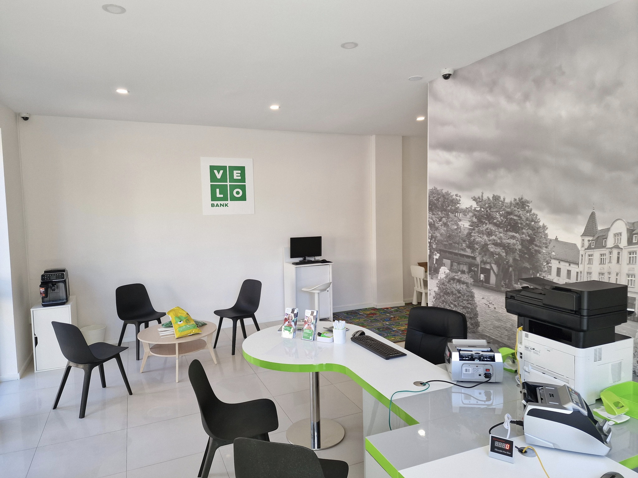 Jasne wnętrze placówki bankowej VeloBank z białymi ścianami, czarnymi krzesłami, biurkiem z zielonym akcentem i sprzętem biurowym, w tle fototapeta z czarno-białym motywem miejskim.