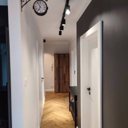 Patrycja Matuszewska P&P Art-Bud - Wnętrze korytarza z drewnianą podłogą ułożoną w jodełkę, białe ściany z dekoracyjnym zegarem na kutym wsporniku, czarne oświetlenie szynowe i ciemnoszara ściana z białymi drzwiami.