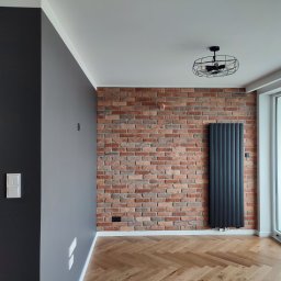 Patrycja Matuszewska P&P Art-Bud - Pokój po odświeżeniu: ściana z cegły klinkierowej, pionowy grzejnik panelowy w kolorze antracytowym, parkiet w jodełkę, ściany pomalowane w odcieniach szarości, nowoczesna lampa sufitowa w stylu...