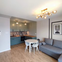 Patrycja Matuszewska P&P Art-Bud - Jasne wnętrze mieszkania po remoncie: zielona zabudowa kuchenna z drewnianym blatem, stół z białymi krzesłami, szara sofa, parkiet i nowoczesna lampa sufitowa.