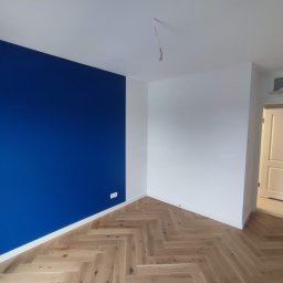 Patrycja Matuszewska P&P Art-Bud - Pomalowane na biało i granatowo wnętrze pokoju z parkietem ułożonym w jodełkę, widoczne gniazdka elektryczne na ścianie i otwarte drzwi do innego pomieszczenia.