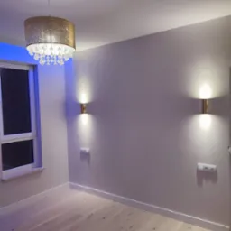 Jasne panele podłogowe, szare ściany z dwoma złotymi kinkietami i gniazdkami elektrycznymi, okno z niebieskim podświetleniem oraz złoty żyrandol z kryształkami w pustym pokoju.