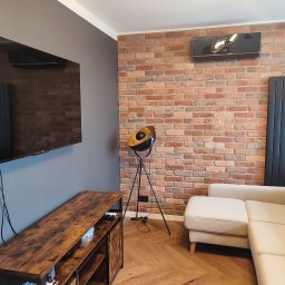 Patrycja Matuszewska P&P Art-Bud - Salon z ceglaną ścianą, beżową sofą narożną, telewizorem na szarej ścianie i drewnianą szafką RTV, oświetlony lampą na trójnogu.