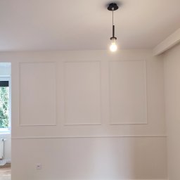 Patrycja Matuszewska P&P Art-Bud - Jasne wnętrze z drewnianą podłogą, białe ściany z dekoracyjnymi ramkami i listwami, nowoczesna lampa wisząca z żarówką, widok na okno z zielenią na zewnątrz.