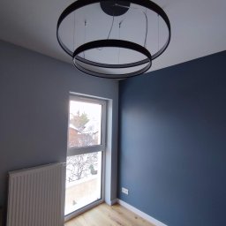 Patrycja Matuszewska P&P Art-Bud - Wnętrze pokoju po odświeżeniu: szare ściany, granatowa akcentująca ściana, drewniana podłoga, biały grzejnik pod oknem z widokiem na zimowy krajobraz, nowoczesna dwuobręczowa lampa sufitowa.