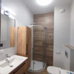 Nowoczesna łazienka z kabiną prysznicową narożną, umywalką z szafką z drewnianymi frontami, toaletą i pralką, wykończona płytkami w odcieniach bieli i drewna.