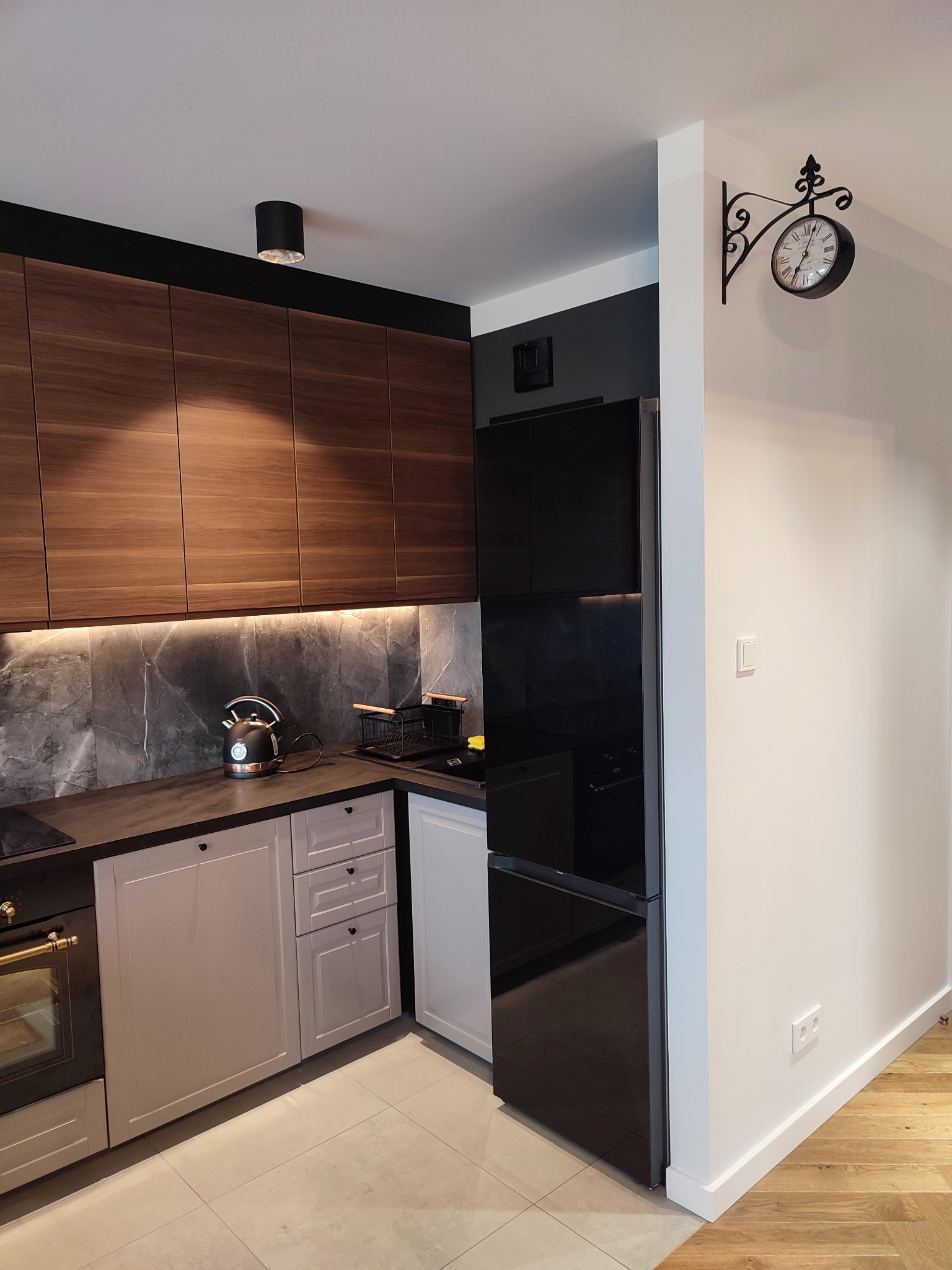 Nowoczesna kuchnia w odcieniach brązu, szarości i bieli z drewnianymi szafkami, marmurowym backsplash, czarnym czajnikiem i zegarem w stylu retro na ścianie.