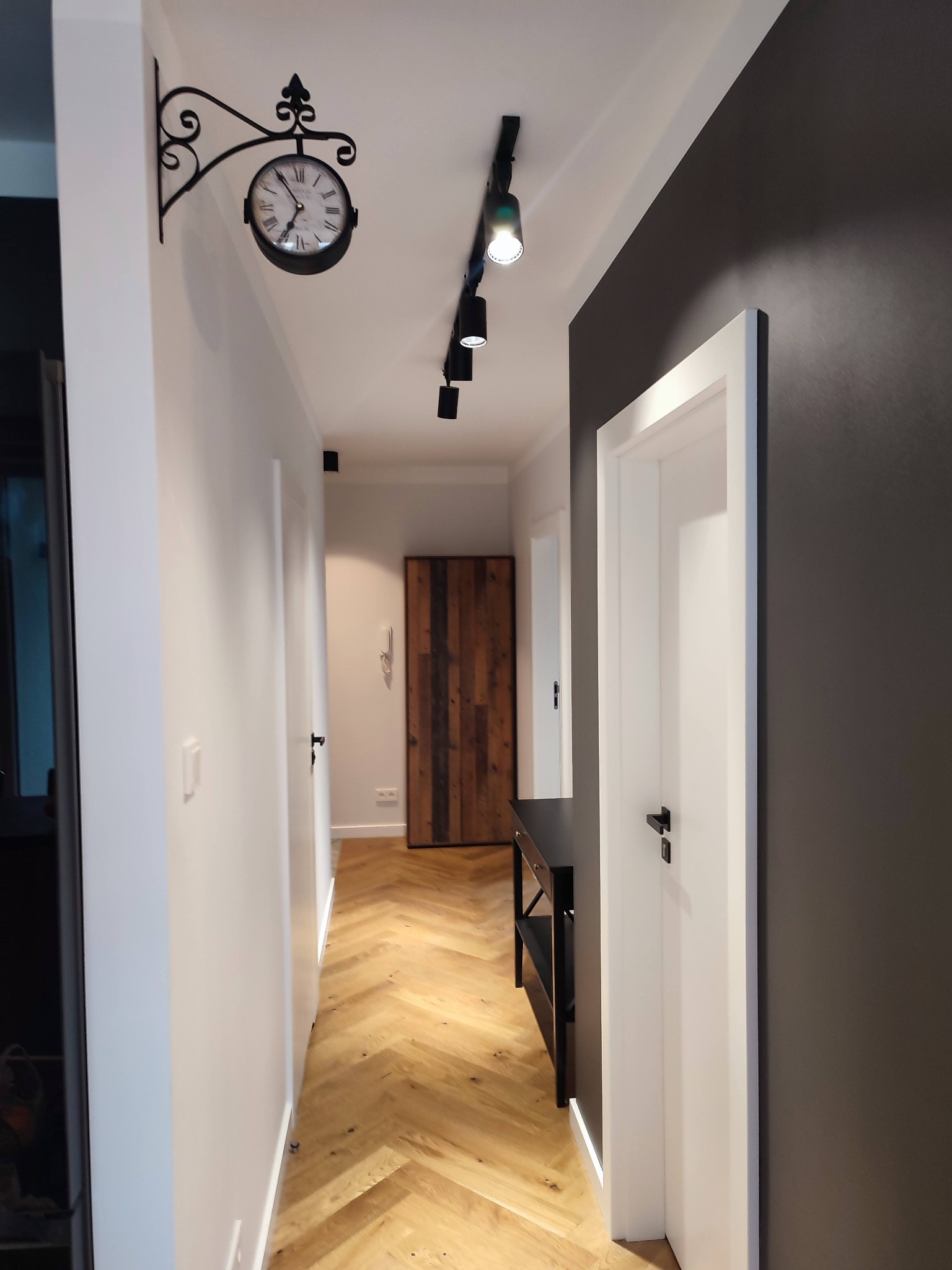 Wnętrze korytarza z drewnianą podłogą ułożoną w jodełkę, białe ściany z dekoracyjnym zegarem na kutym wsporniku, czarne oświetlenie szynowe i ciemnoszara ściana z białymi drzwiami.