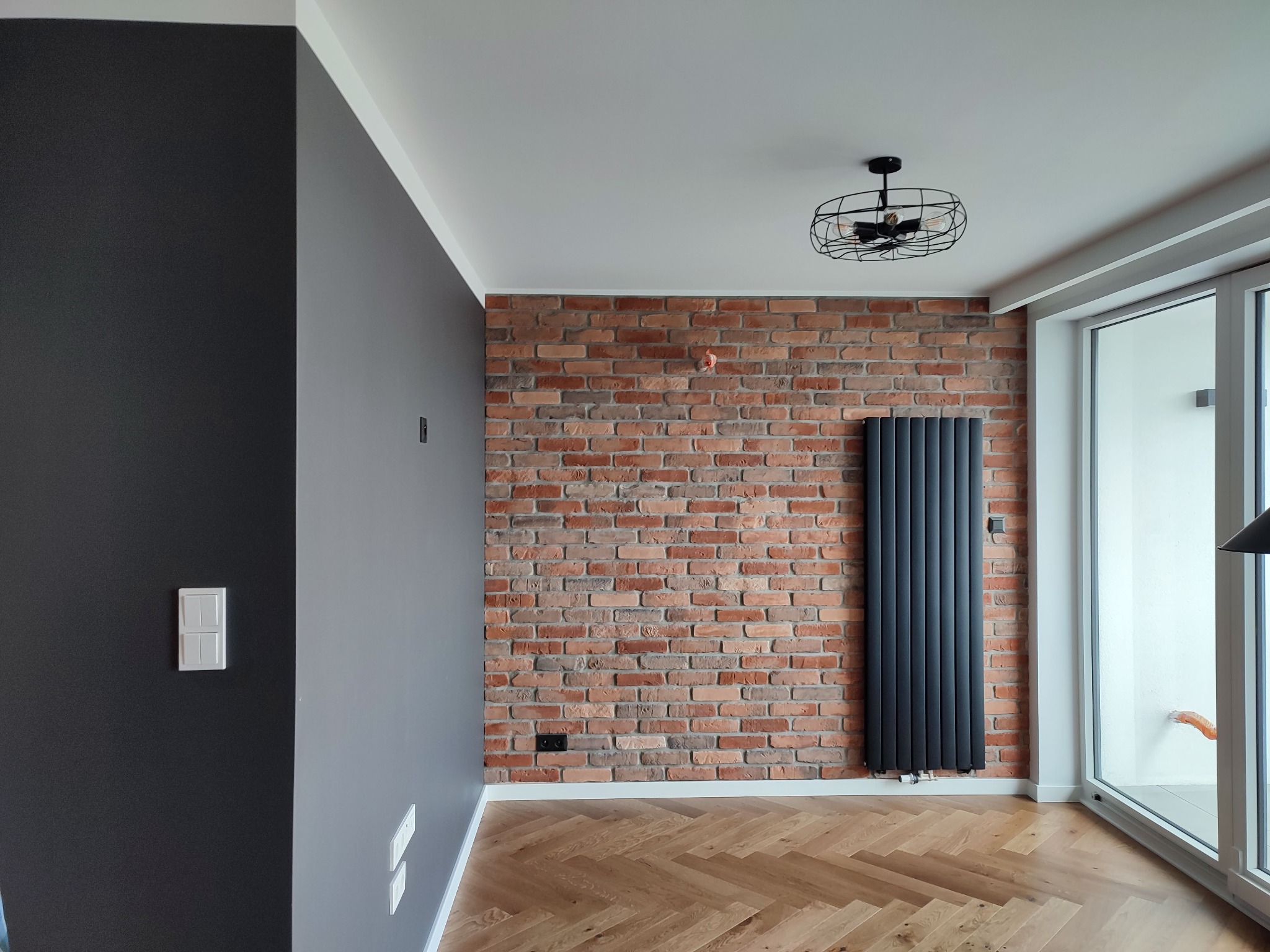 Pokój po odświeżeniu: ściana z cegły klinkierowej, pionowy grzejnik panelowy w kolorze antracytowym, parkiet w jodełkę, ściany pomalowane w odcieniach szarości, nowoczesna lampa sufitowa w stylu...