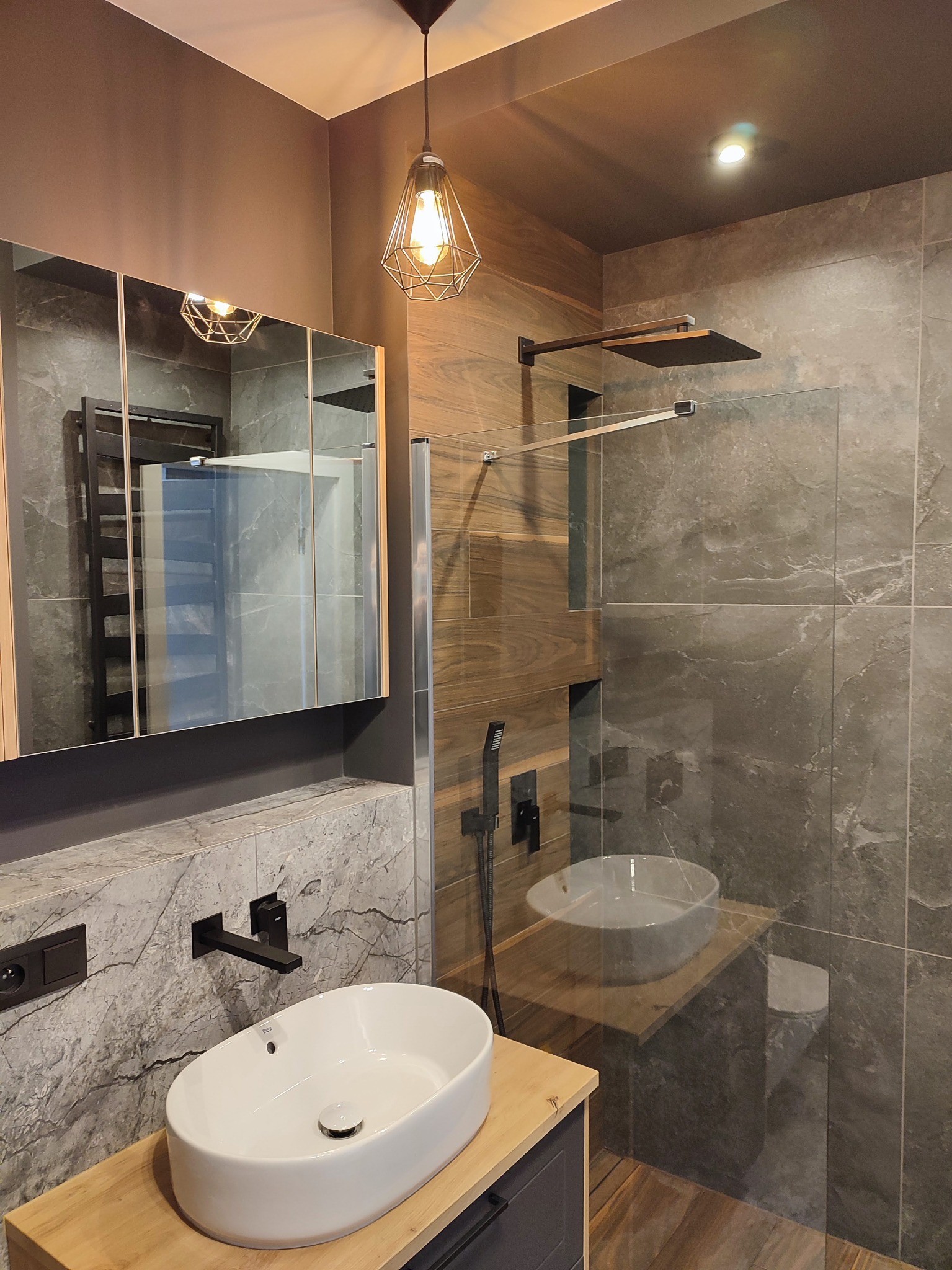 Nowoczesna łazienka z szarymi płytkami imitującymi marmur, przeszkloną kabiną prysznicową typu walk-in, białą umywalką nablatową na drewnianym blacie i czarną armaturą.