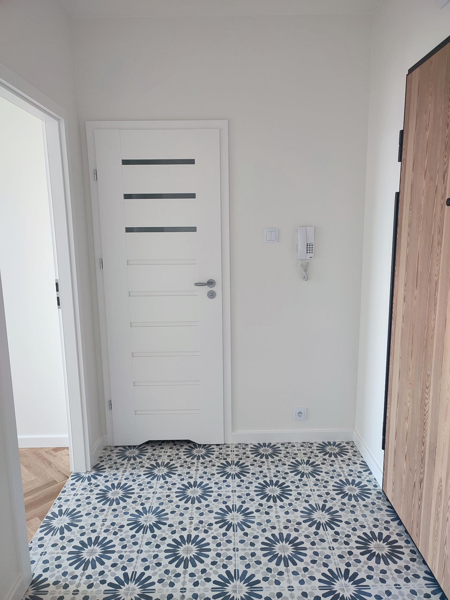 Wykończony przedpokój z wzorzystą, niebiesko-szarą podłogą z płytek, białe ściany, białe drzwi wewnętrzne z poziomymi przeszkleniami, drewniane drzwi wejściowe i domofon na ścianie.
