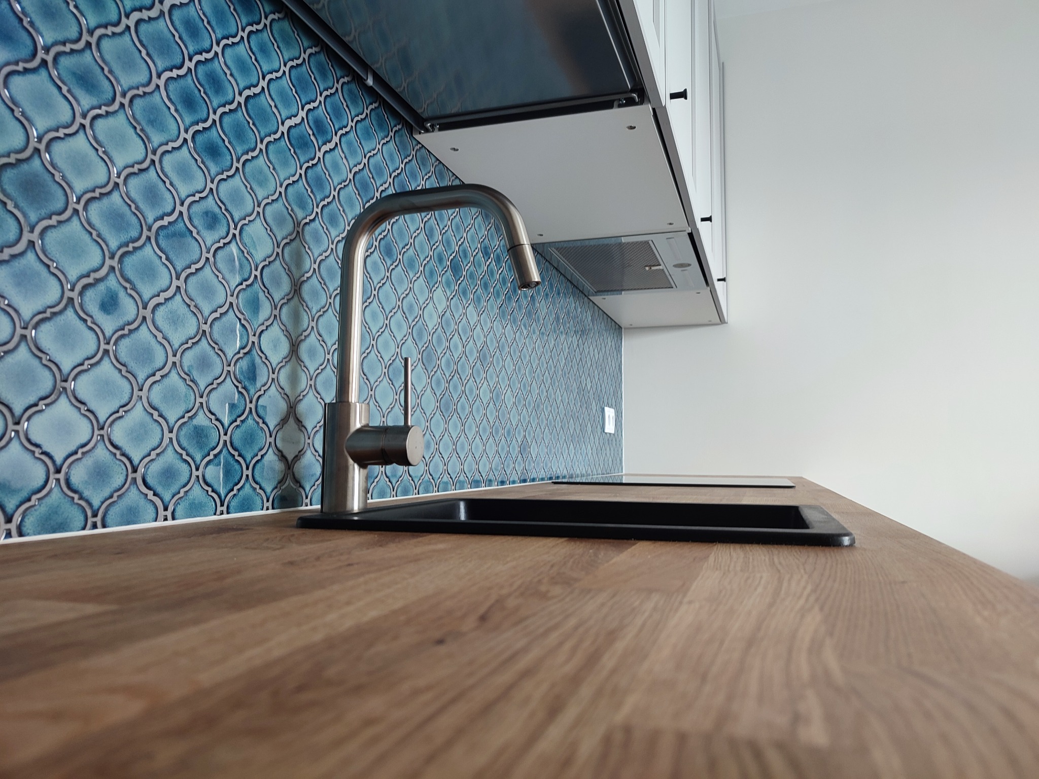 Błękitne mozaikowe płytki nad blatem kuchennym z drewnianym wzorem, widoczny chromowany kran i czarny zlew, ujęcie z niskiej perspektywy.