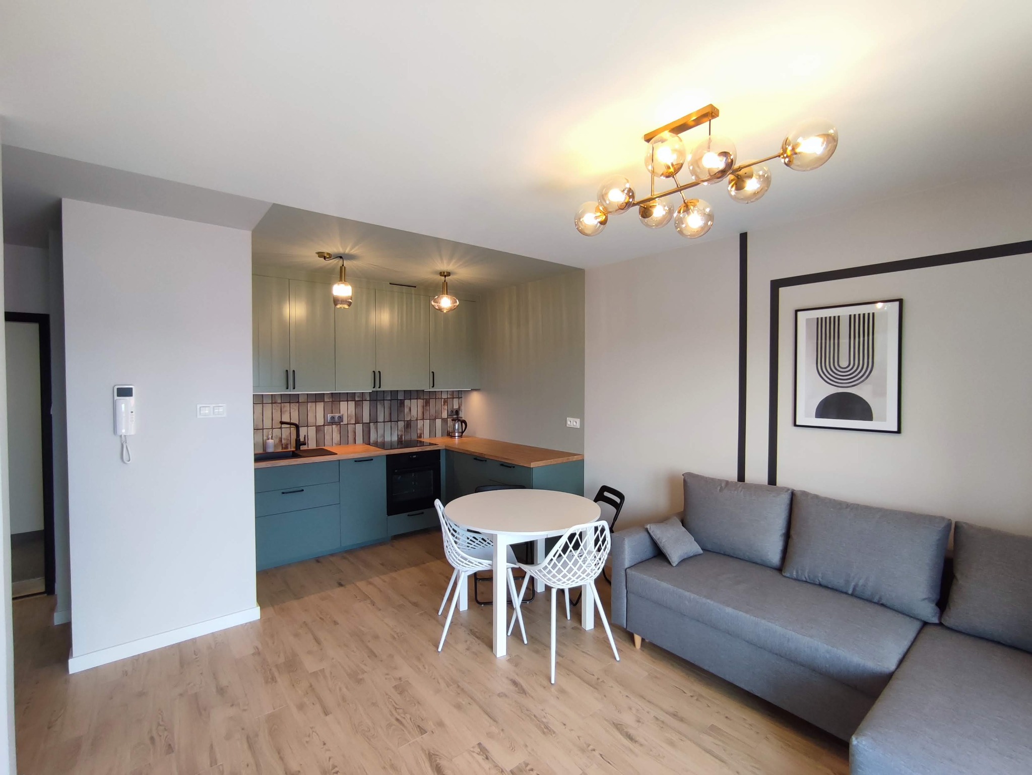 Jasne wnętrze mieszkania po remoncie: zielona zabudowa kuchenna z drewnianym blatem, stół z białymi krzesłami, szara sofa, parkiet i nowoczesna lampa sufitowa.