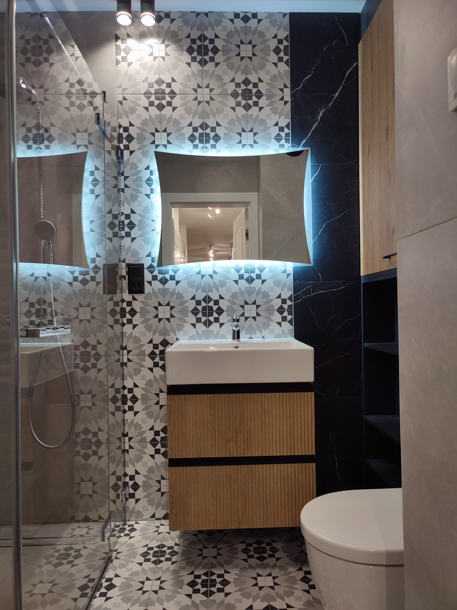 Nowoczesna łazienka z kabiną prysznicową, umywalką na szafce z drewnianymi lamelami, podświetlanym lustrem i płytkami w geometryczne wzory, akcent czarnego marmuru na ścianie.