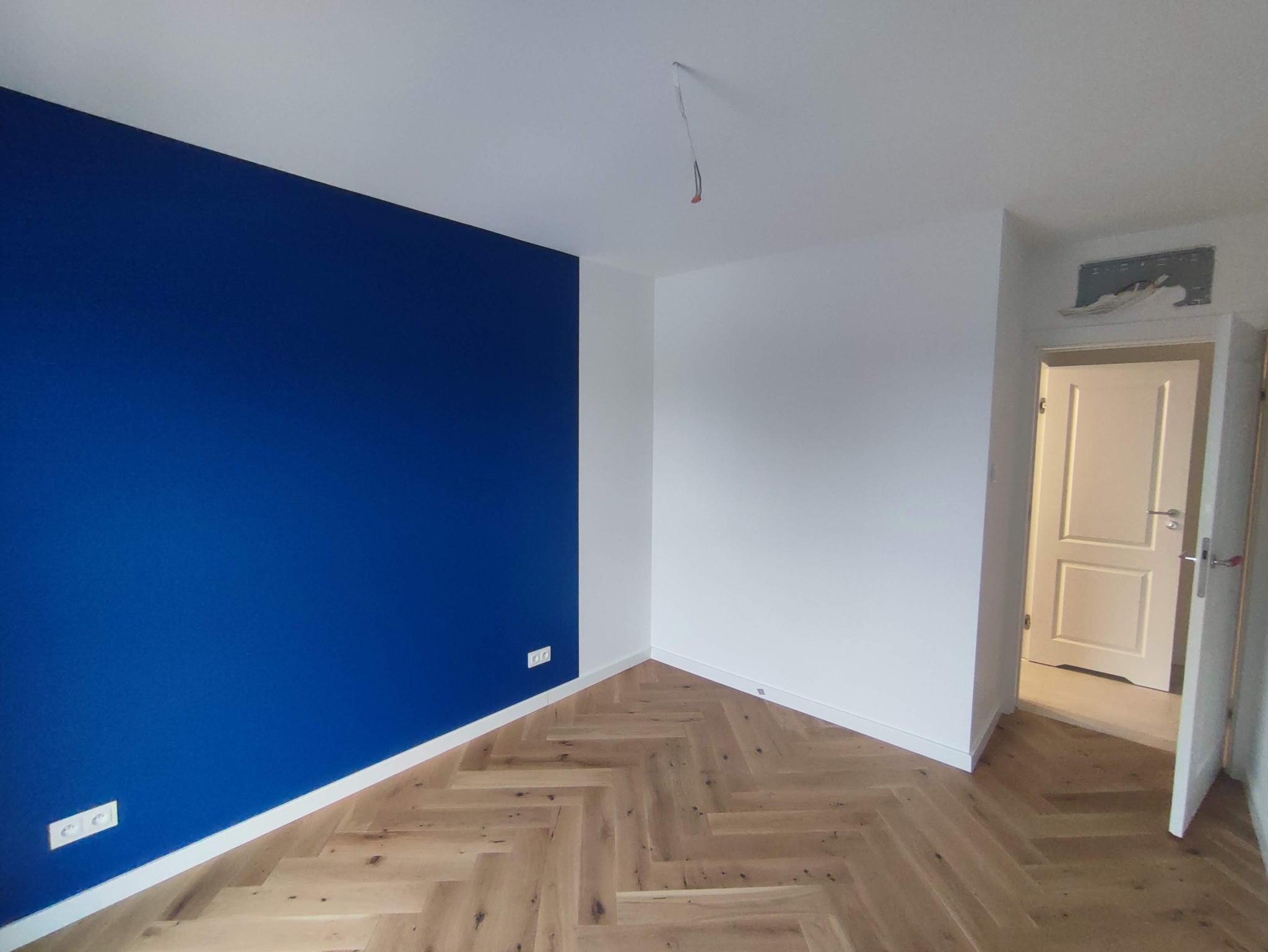 Pomalowane na biało i granatowo wnętrze pokoju z parkietem ułożonym w jodełkę, widoczne gniazdka elektryczne na ścianie i otwarte drzwi do innego pomieszczenia.