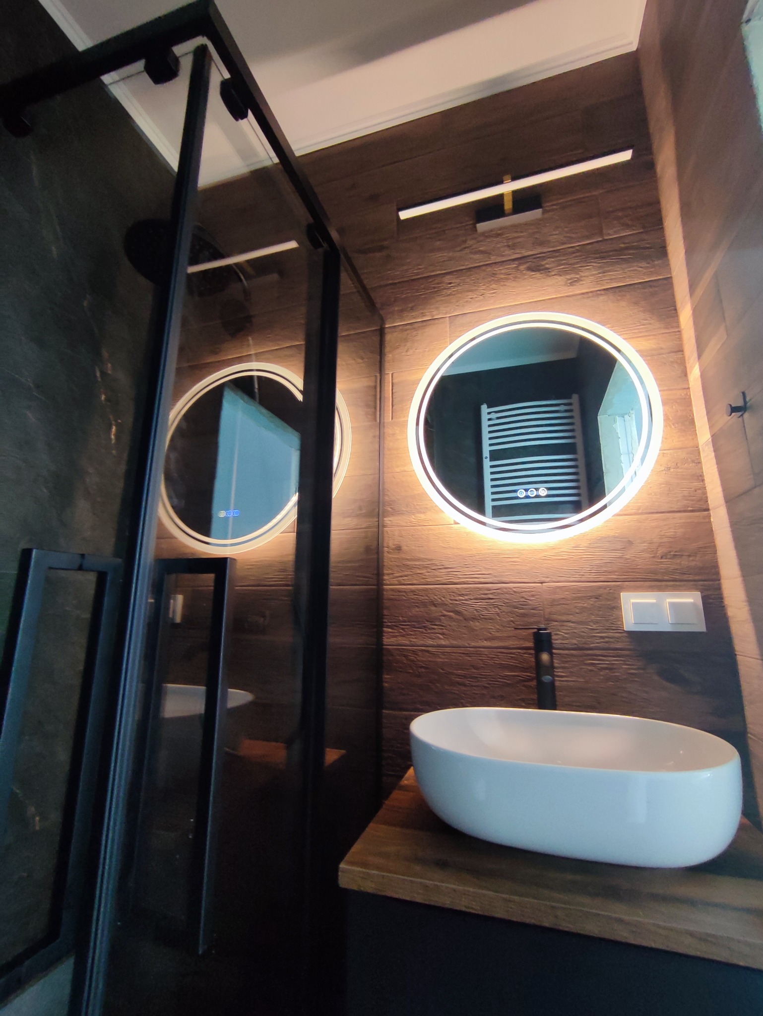 Nowoczesna łazienka z drewnopodobnymi płytkami, owalną umywalką nablatową, podświetlanymi okrągłymi lustrami i czarną kabiną prysznicową typu walk-in.
