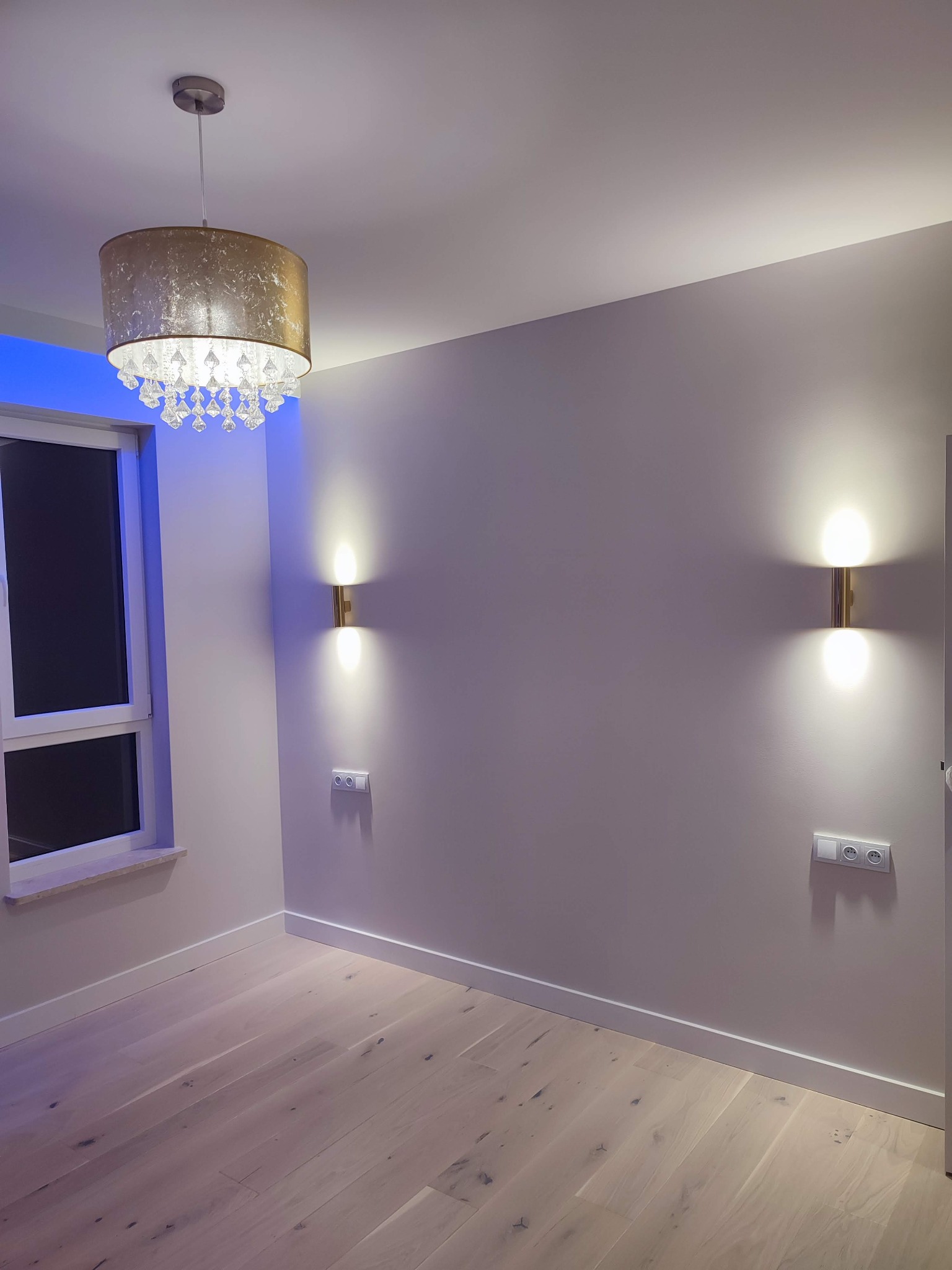 Jasne panele podłogowe, szare ściany z dwoma złotymi kinkietami i gniazdkami elektrycznymi, okno z niebieskim podświetleniem oraz złoty żyrandol z kryształkami w pustym pokoju.
