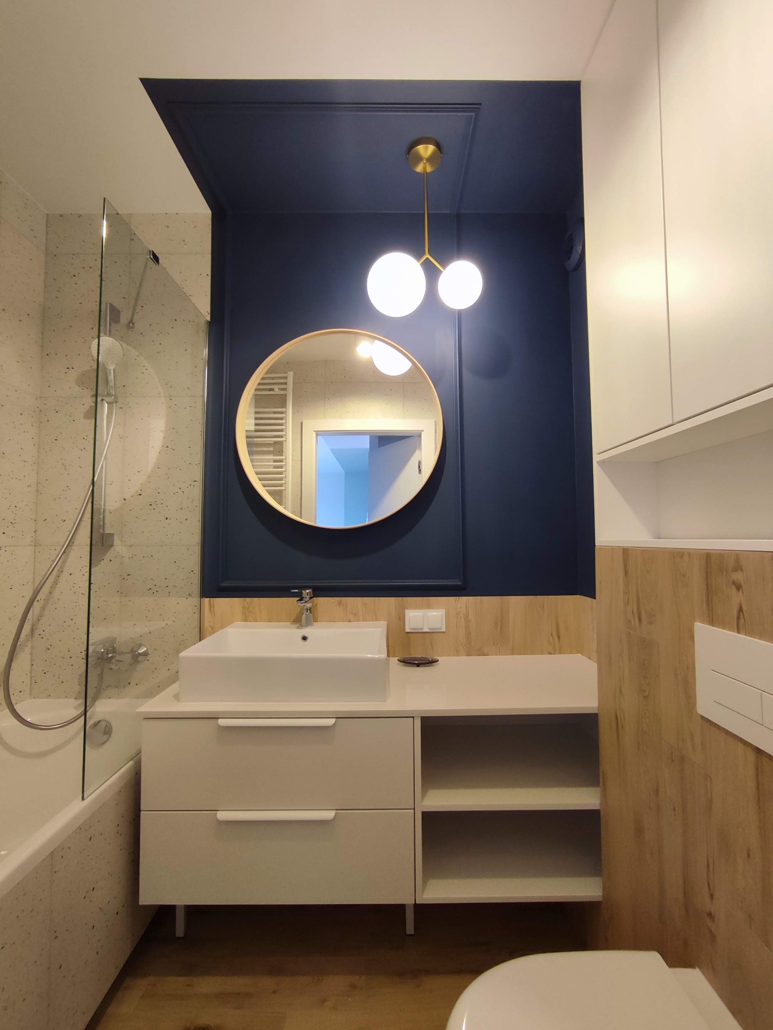 Nowoczesna łazienka z białą umywalką nablatową, okrągłym lustrem w drewnianej ramie na tle granatowej ściany z dekoracyjną listwą, oraz złotą lampą z dwoma kulami, widok na kabinę prysznicową...