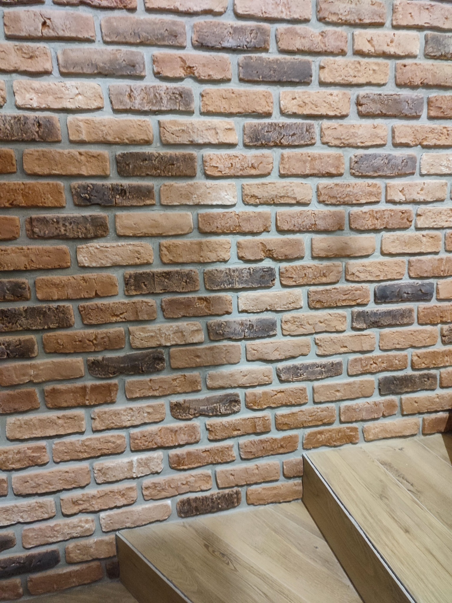 Ściana wyłożona dekoracyjną cegłą w odcieniach brązu i szarości, z fragmentem drewnianych schodów w dolnym prawym rogu kadru.