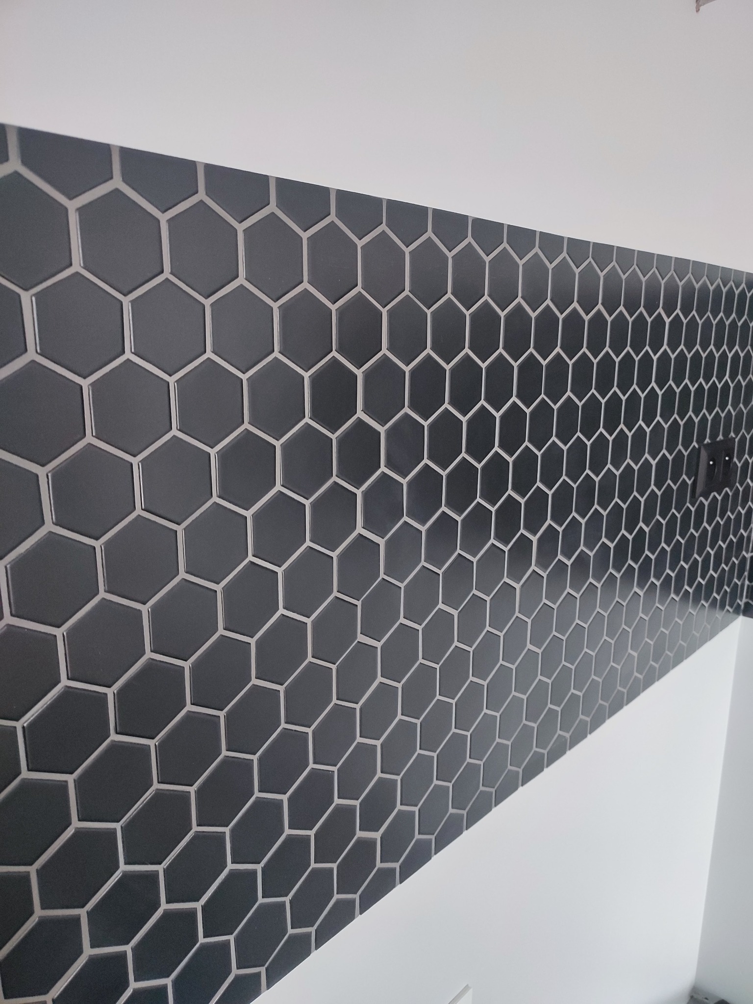 Ściana wyłożona ciemnymi, heksagonalnymi płytkami z jasną fugą, widoczny fragment ściany pomalowanej na biało oraz gniazdko elektryczne.