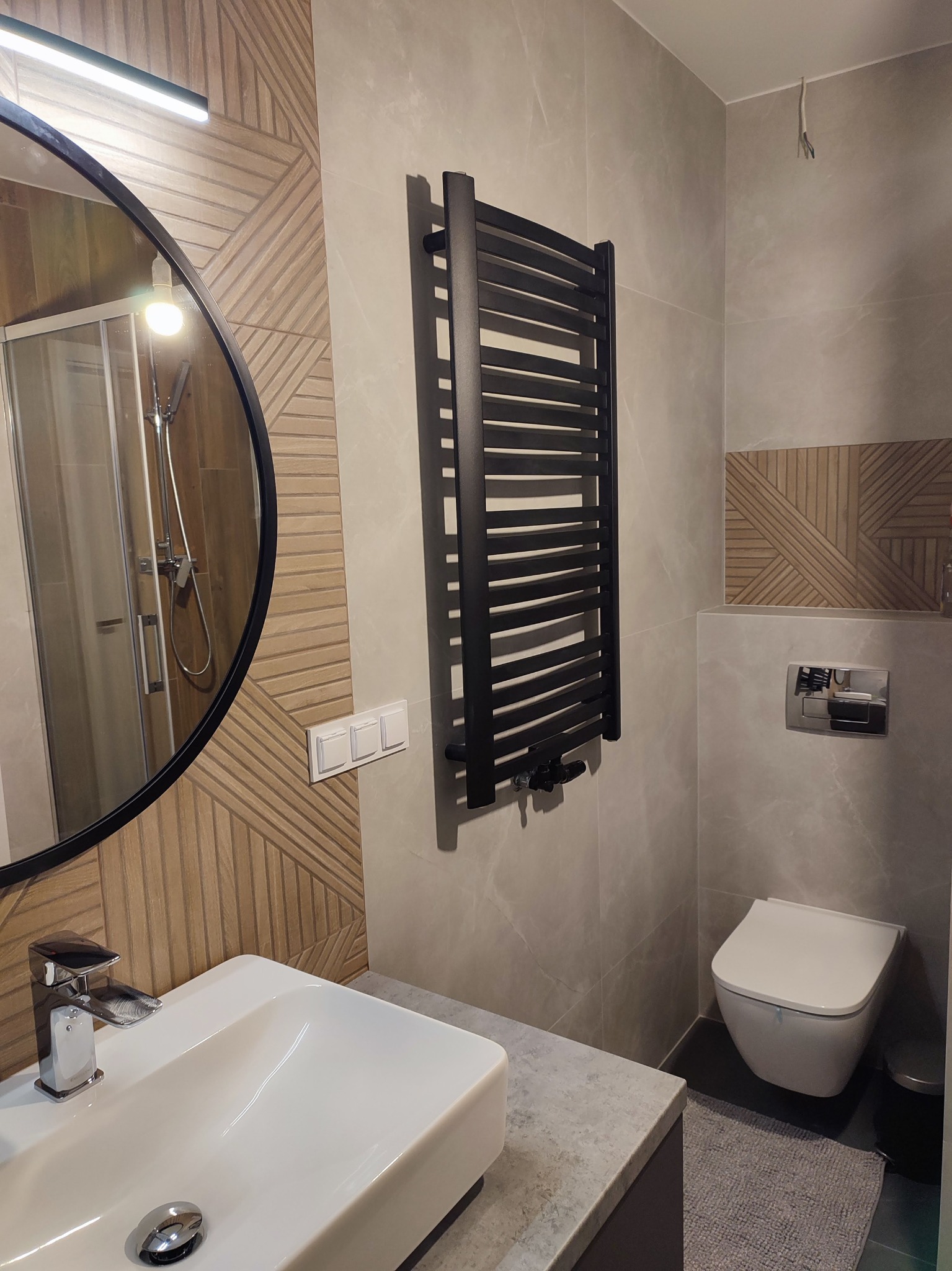 Nowoczesna łazienka z owalnym lustrem, grzejnikiem drabinkowym w kolorze czarnym, białą umywalką nablatową i toaletą podwieszaną, wykończona płytkami w odcieniach szarości i drewna.