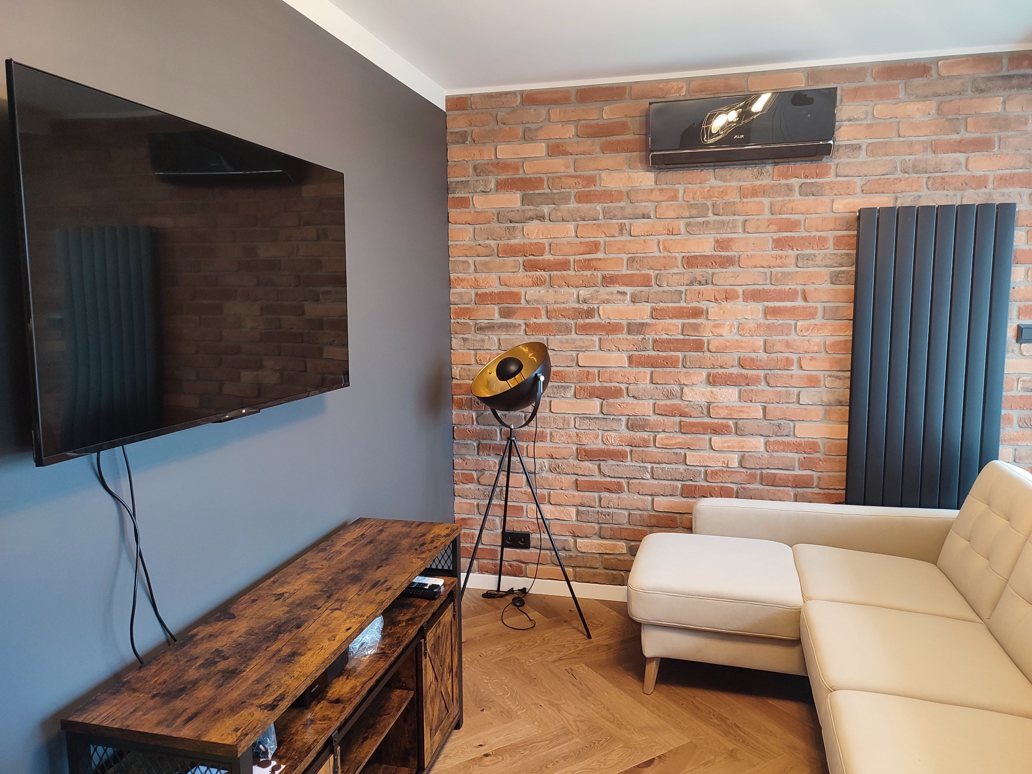 Salon z ceglaną ścianą, beżową sofą narożną, telewizorem na szarej ścianie i drewnianą szafką RTV, oświetlony lampą na trójnogu.