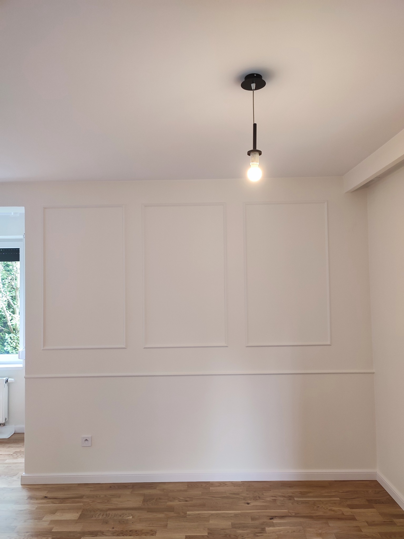 Jasne wnętrze z drewnianą podłogą, białe ściany z dekoracyjnymi ramkami i listwami, nowoczesna lampa wisząca z żarówką, widok na okno z zielenią na zewnątrz.