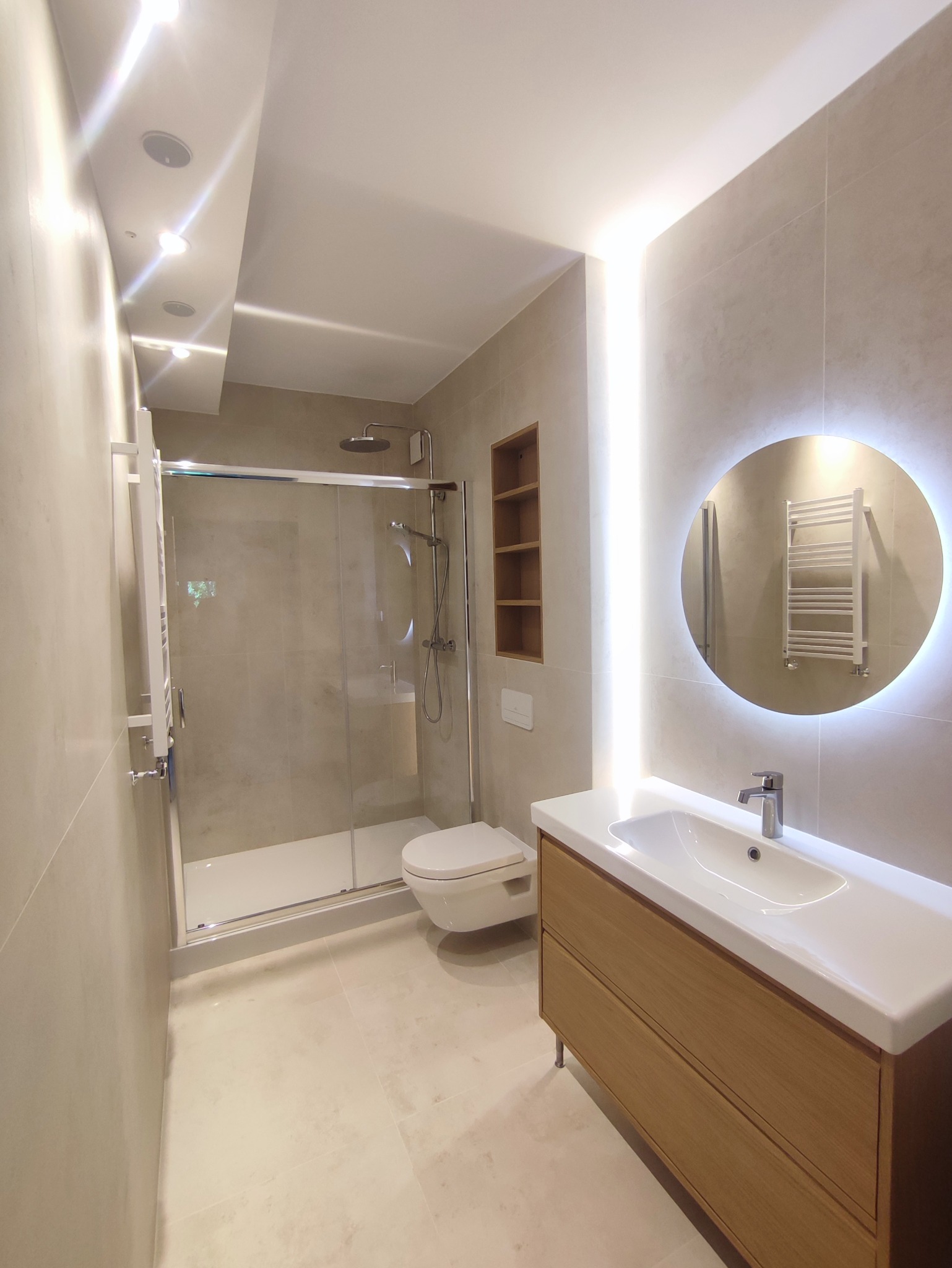 Nowoczesna łazienka z kabiną prysznicową, podwieszaną toaletą, szafką z umywalką z drewnianymi frontami i okrągłym lustrem z podświetleniem LED.