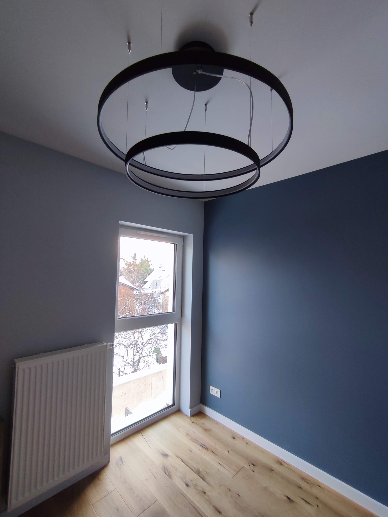Wnętrze pokoju po odświeżeniu: szare ściany, granatowa akcentująca ściana, drewniana podłoga, biały grzejnik pod oknem z widokiem na zimowy krajobraz, nowoczesna dwuobręczowa lampa sufitowa.