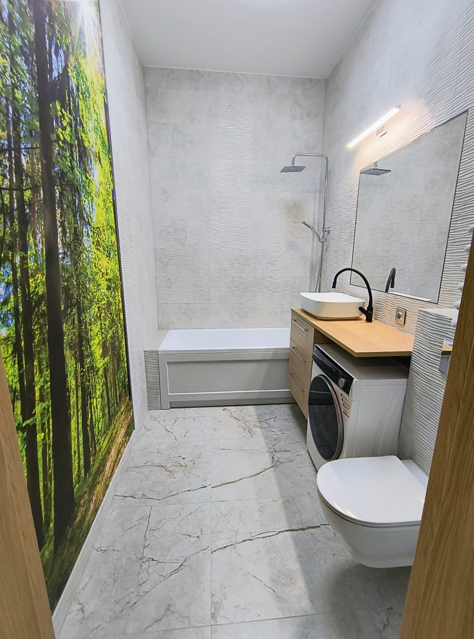 Nowoczesna łazienka z fototapetą leśną, umywalką nablatową na drewnianym blacie, pralką pod blatem i wiszącą toaletą; płytki imitujące fale na ścianach i marmurowe na podłodze.