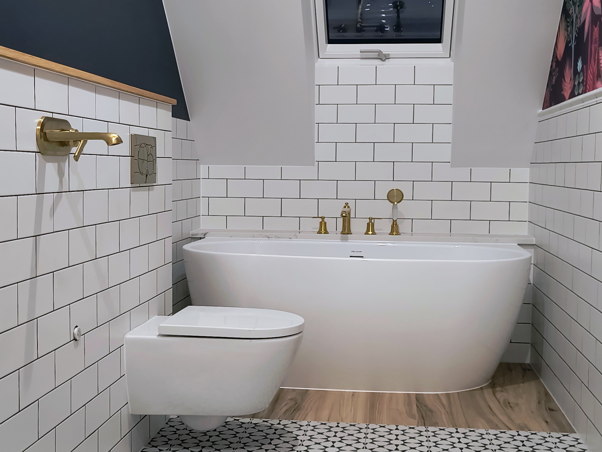 Nowoczesna łazienka na poddaszu z białą wolnostojącą wanną, wiszącą toaletą i złotą armaturą na tle białych płytek ułożonych w cegiełkę, z oknem dachowym i fragmentem dekoracyjnej tapety.