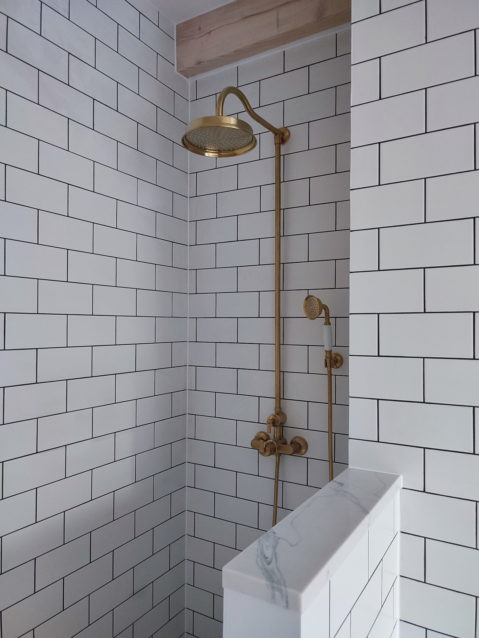 Nowoczesna kabina prysznicowa z białymi, prostokątnymi płytkami i złotą armaturą, wykończona drewnianą belką stropową.