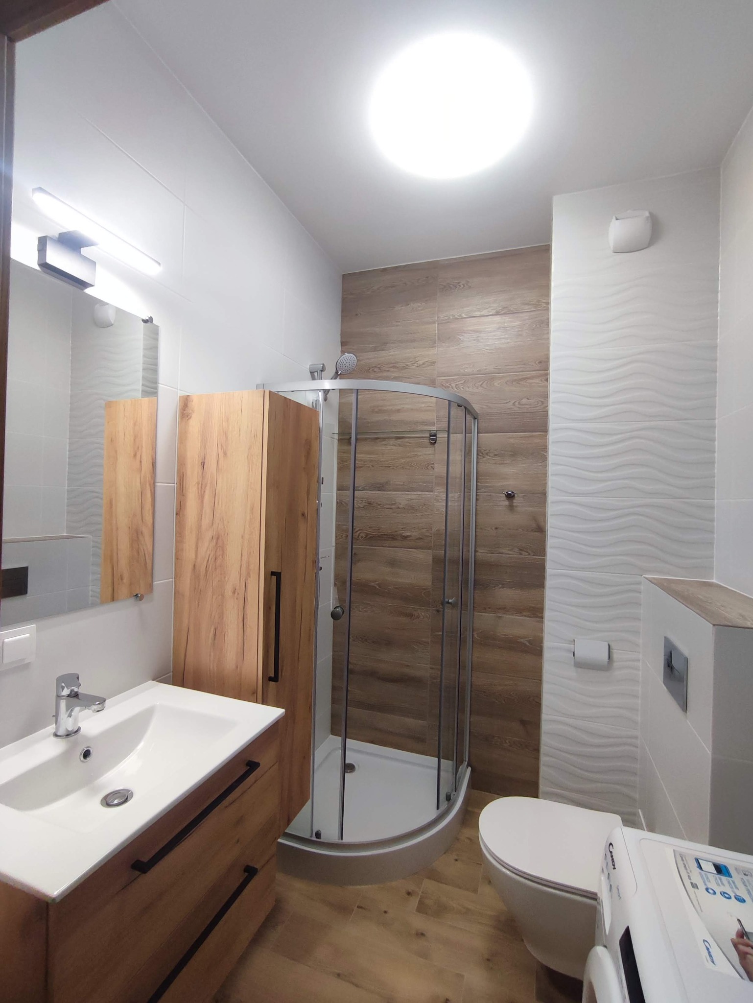 Nowoczesna łazienka z kabiną prysznicową narożną, umywalką z szafką z drewnianymi frontami, toaletą i pralką, wykończona płytkami w odcieniach bieli i drewna.