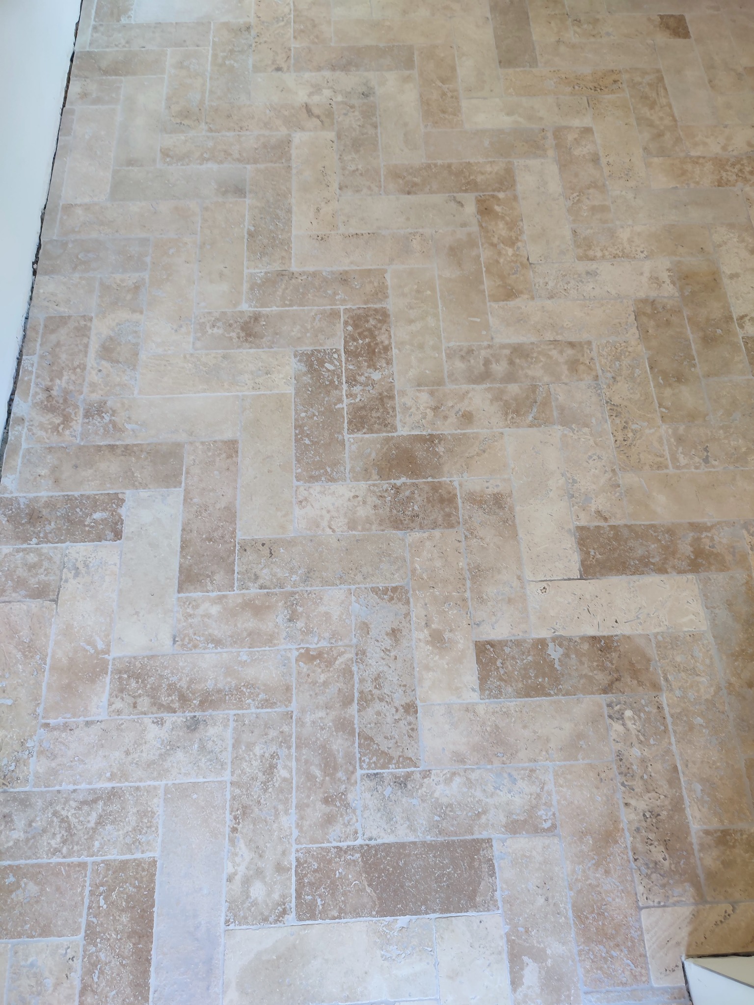 Jasna podłoga z płytek ułożonych w jodełkę, widoczne fugi i niedokończone wykończenie przy ścianie.