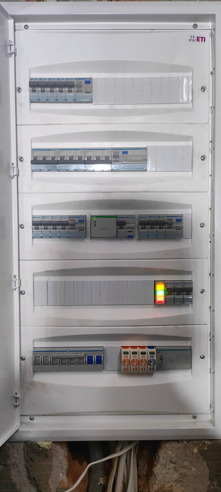 Otwarta skrzynka elektryczna z wieloma bezpiecznikami i wyłącznikami, widoczne kable wychodzące z dołu, jeden wskaźnik świeci na zielono i czerwono.