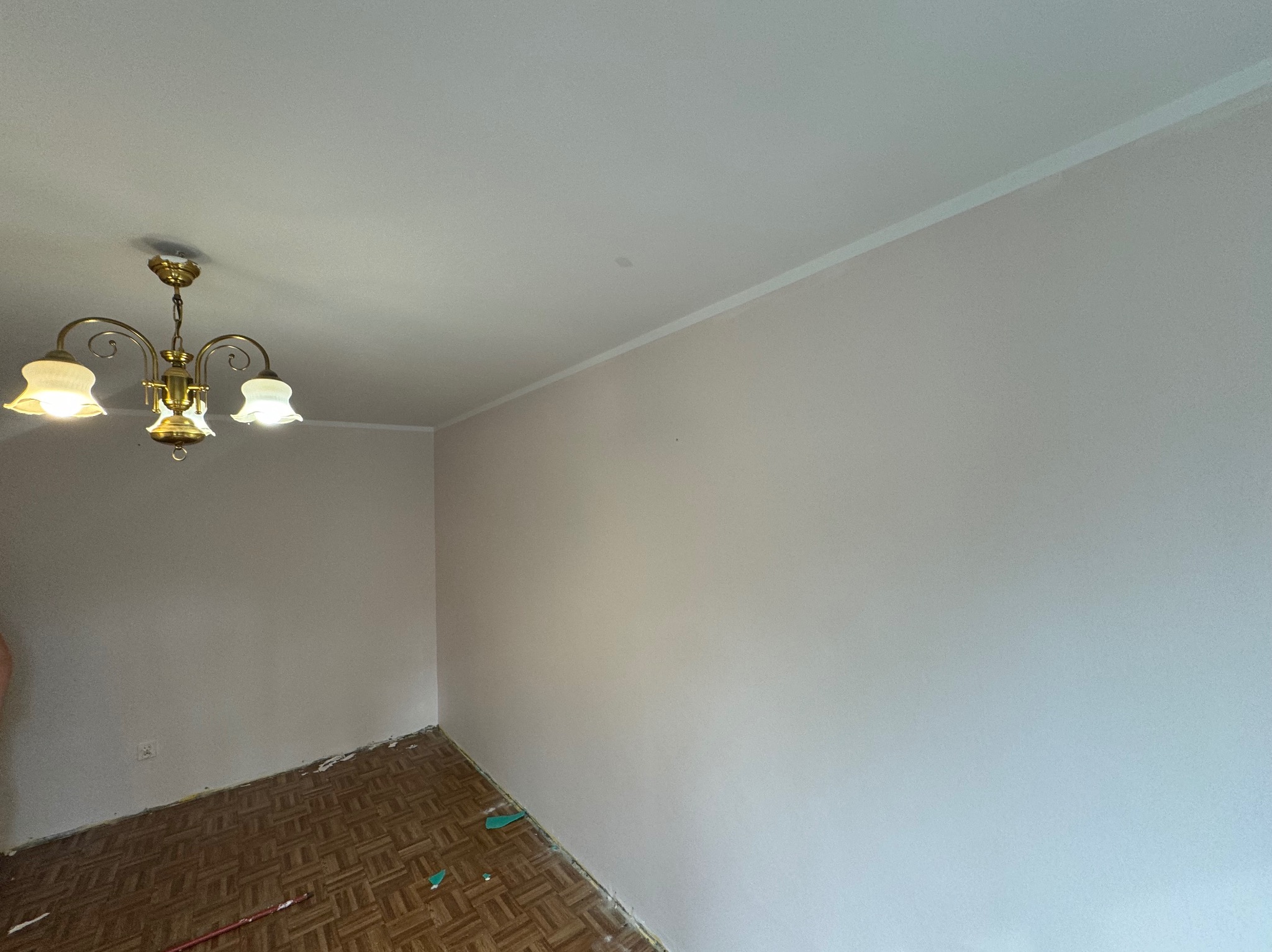 Pomalowane na jasny kolor wnętrze pokoju z parkietem, widoczny złoty żyrandol i ślady po remoncie przy podłodze.