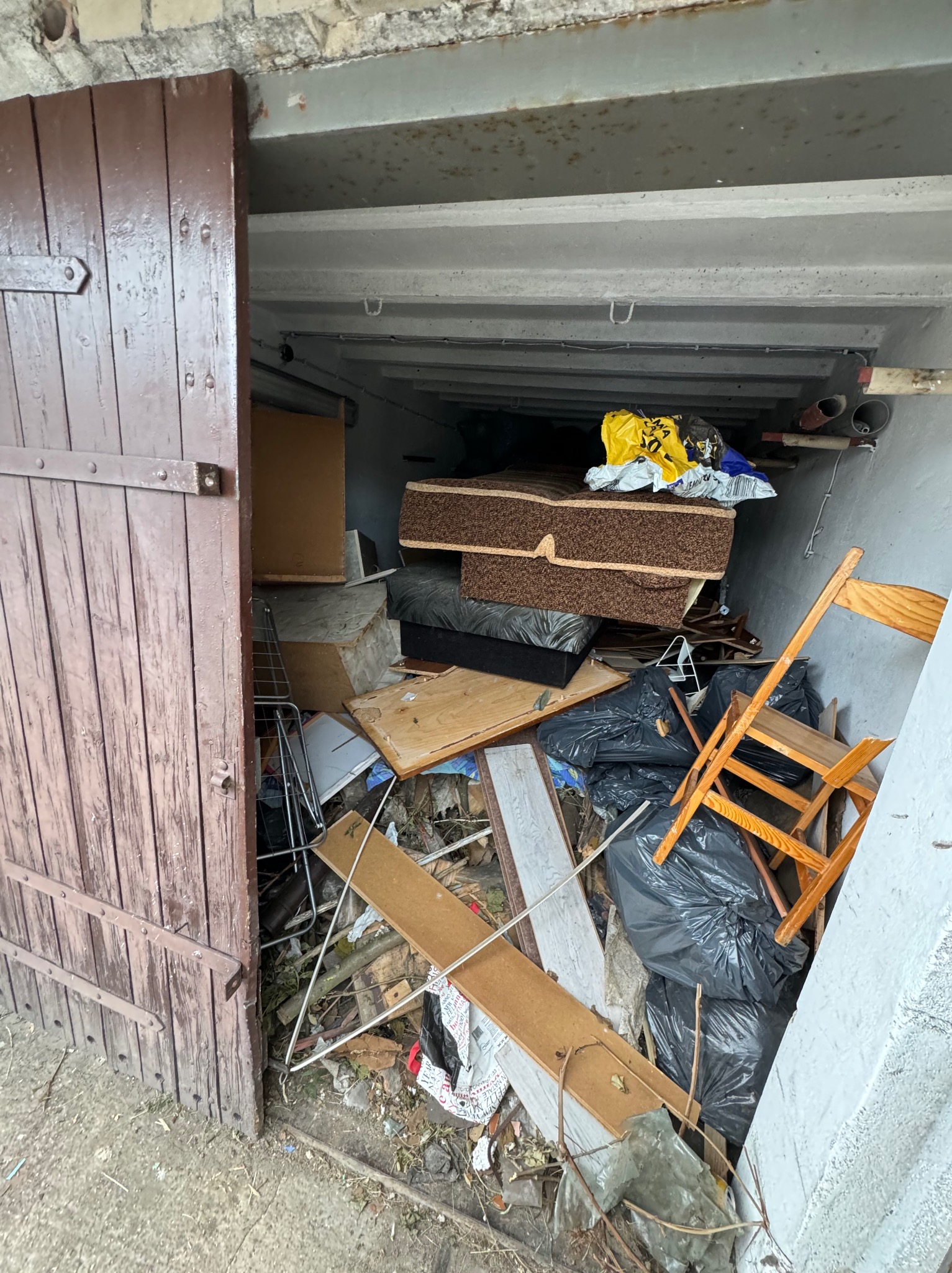 Wnętrze garażu wypełnione różnorodnymi odpadami, w tym meblami, gruzem w czarnych workach i elementami drewnianymi, widoczne przez uchylone, ciemnobrązowe, drewniane wrota.
