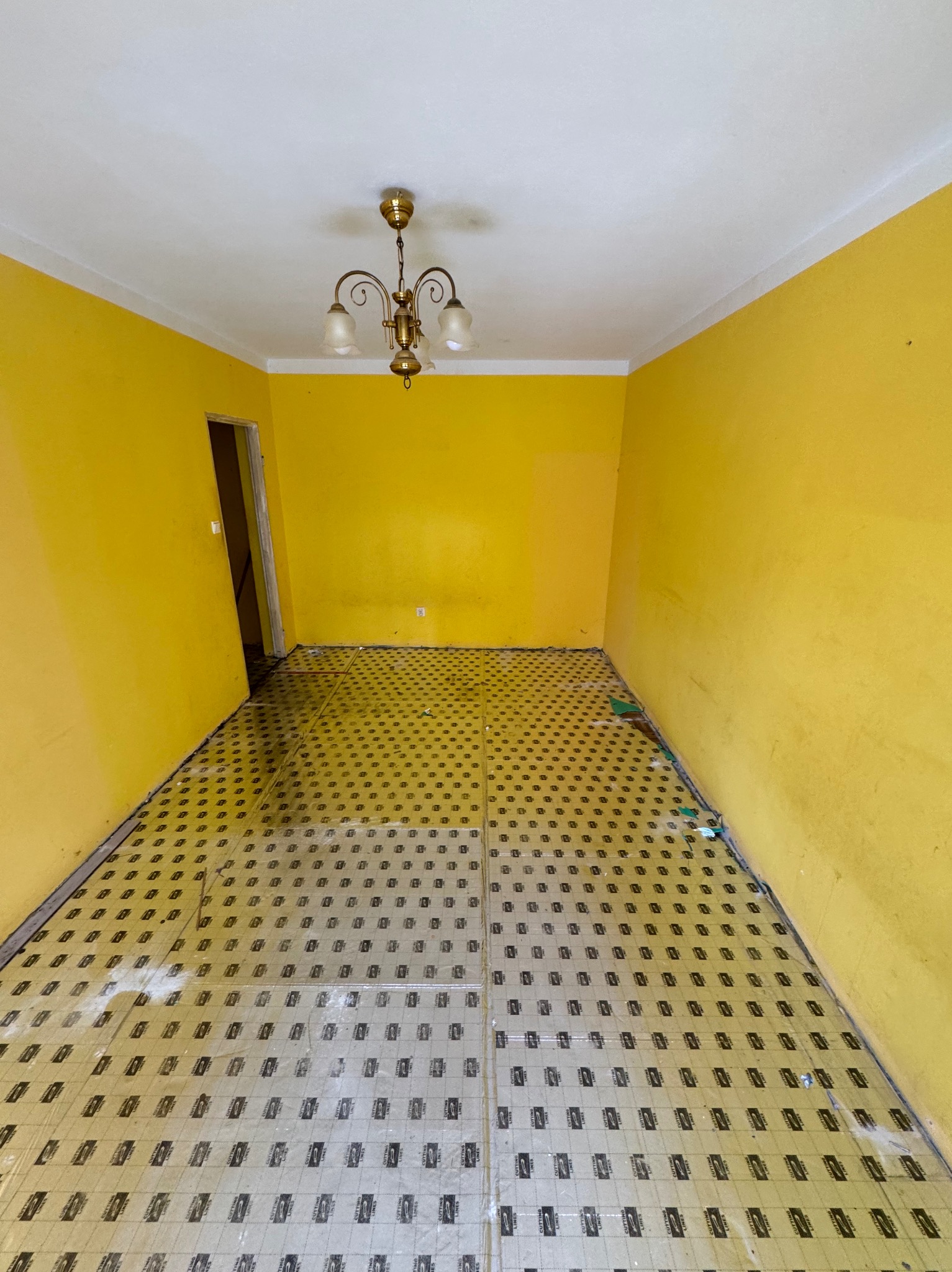 Pokój z żółtymi ścianami w trakcie przygotowania do remontu, widoczne stare płytki podłogowe częściowo przykryte folią ochronną i resztki gruzu, a także wiszący żyrandol.