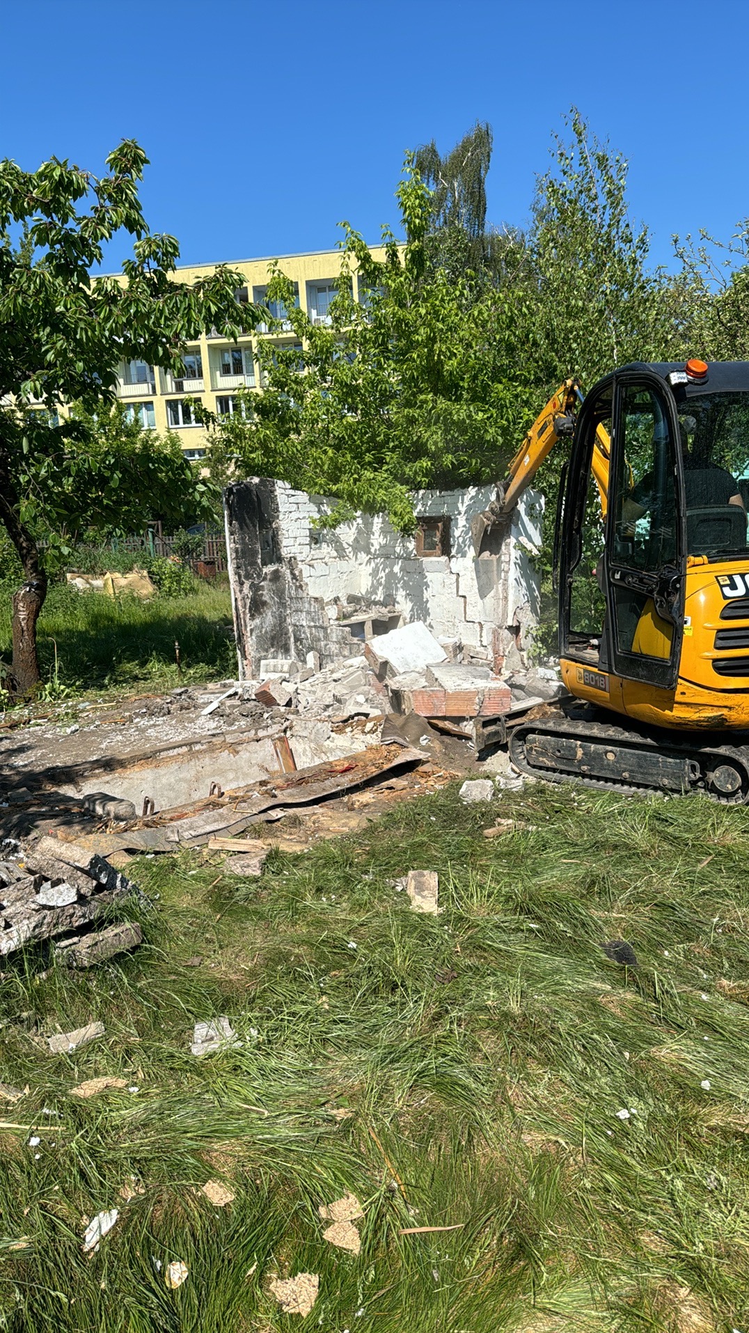 Żółta mini koparka JCB 8018 rozbiera fragment murowanej konstrukcji na zewnątrz, z widocznymi gruzami i otaczającą zielenią oraz budynkiem w tle.