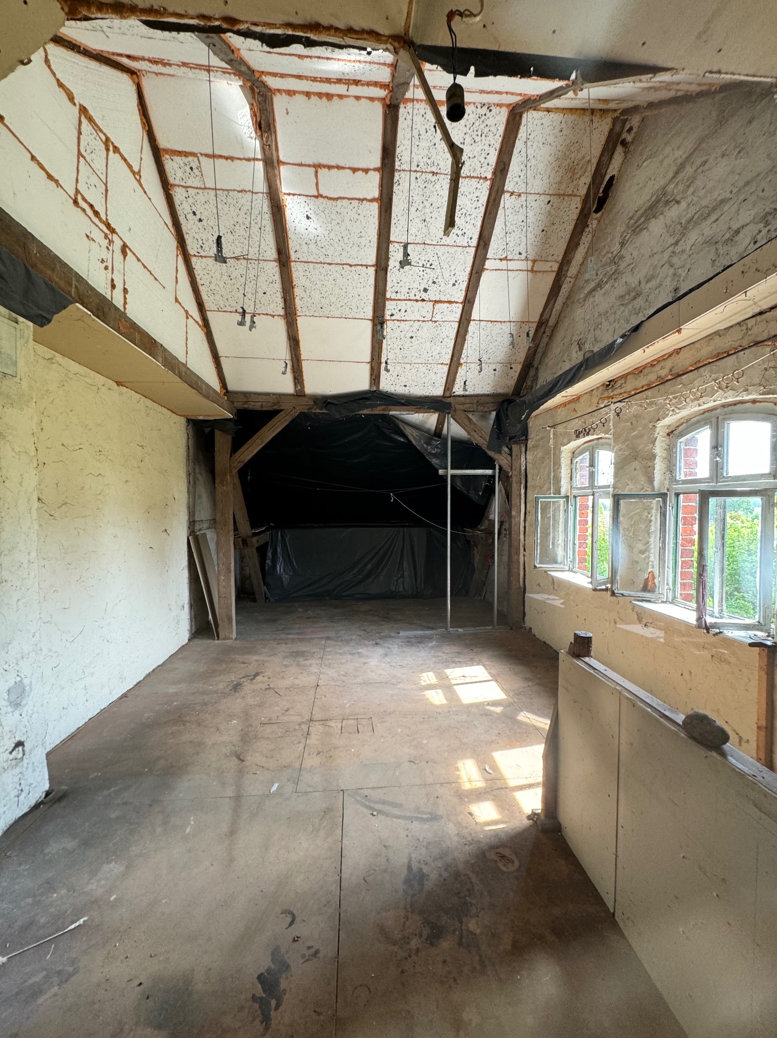 Wnętrze starego budynku w trakcie rozbiórki lub remontu, widoczne odsłonięte belki stropowe, ocieplenie dachu, fragmenty ścian z oknami, podłoga z widocznymi śladami prac.
