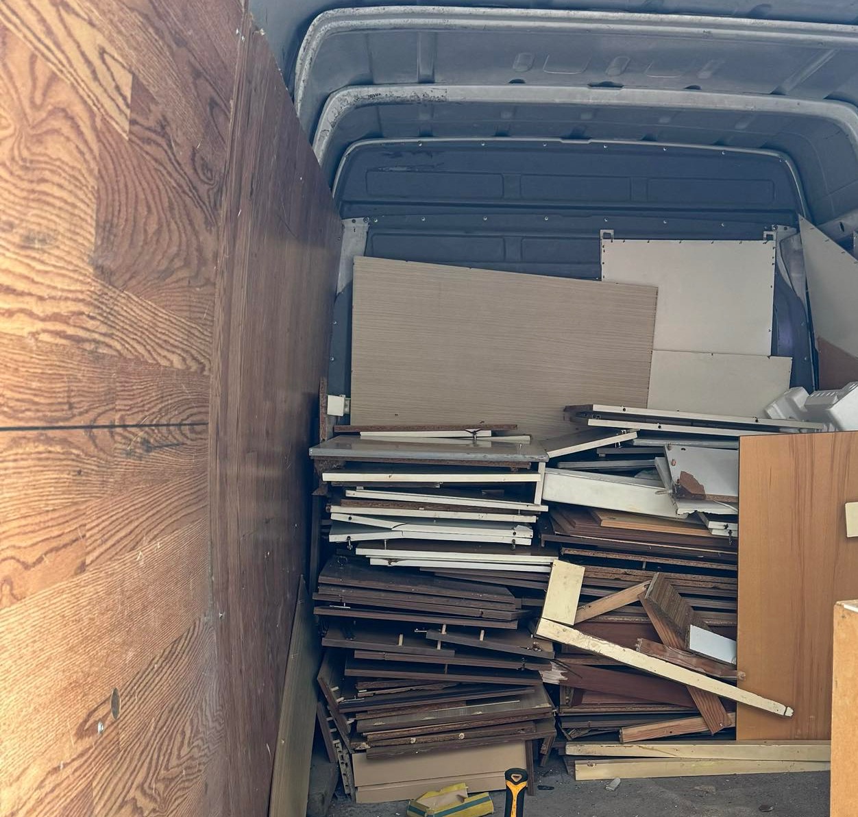 Wnętrze furgonetki wypełnione stertą różnorodnych elementów drewnianych i płyt meblowych, widoczny fragment żółtej miarki na podłodze.